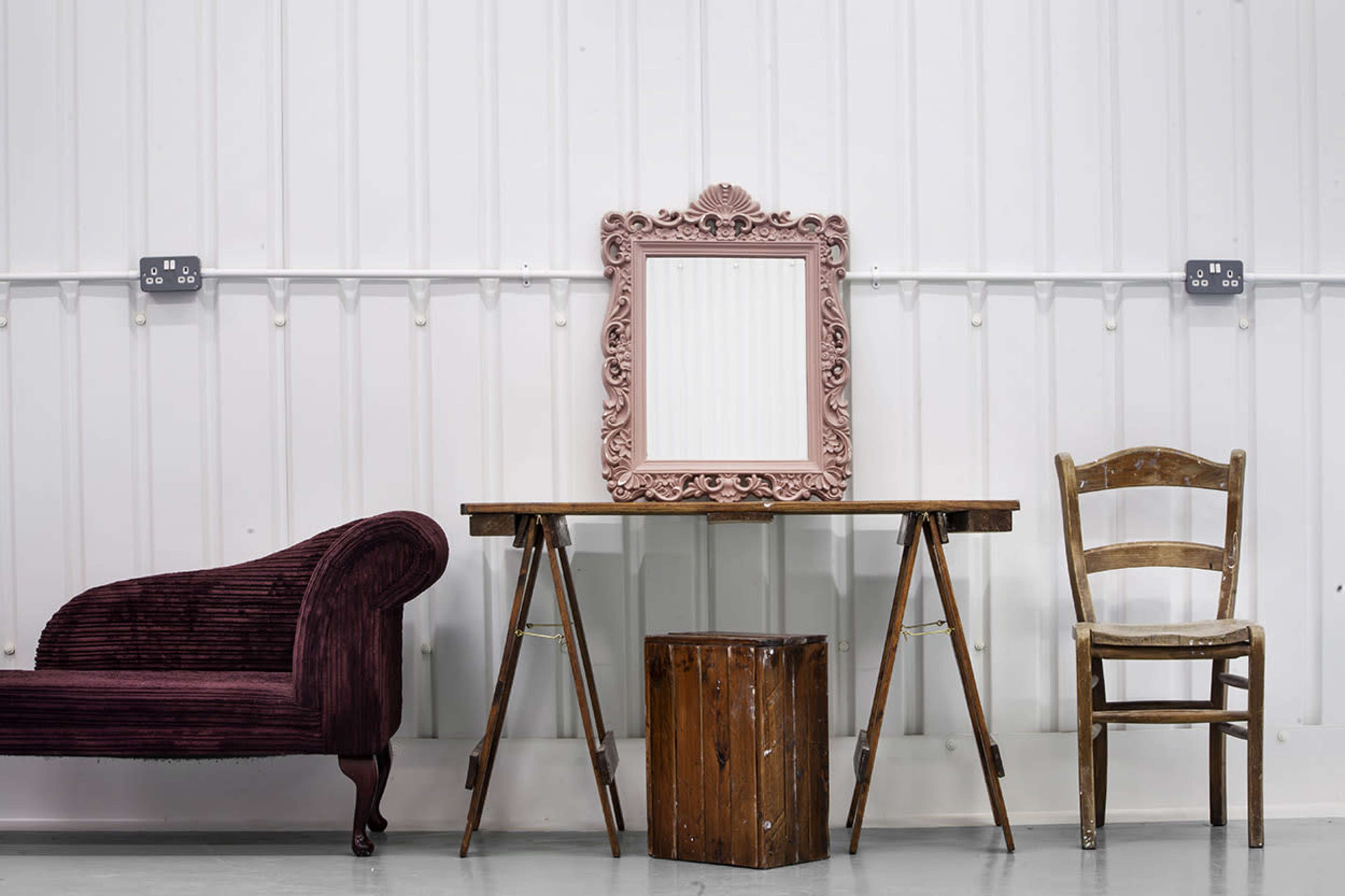 A pink-framed mirror hangs above a wooden table supported by trestles, flanked by a plush couch and a wooden chair against a white wall.