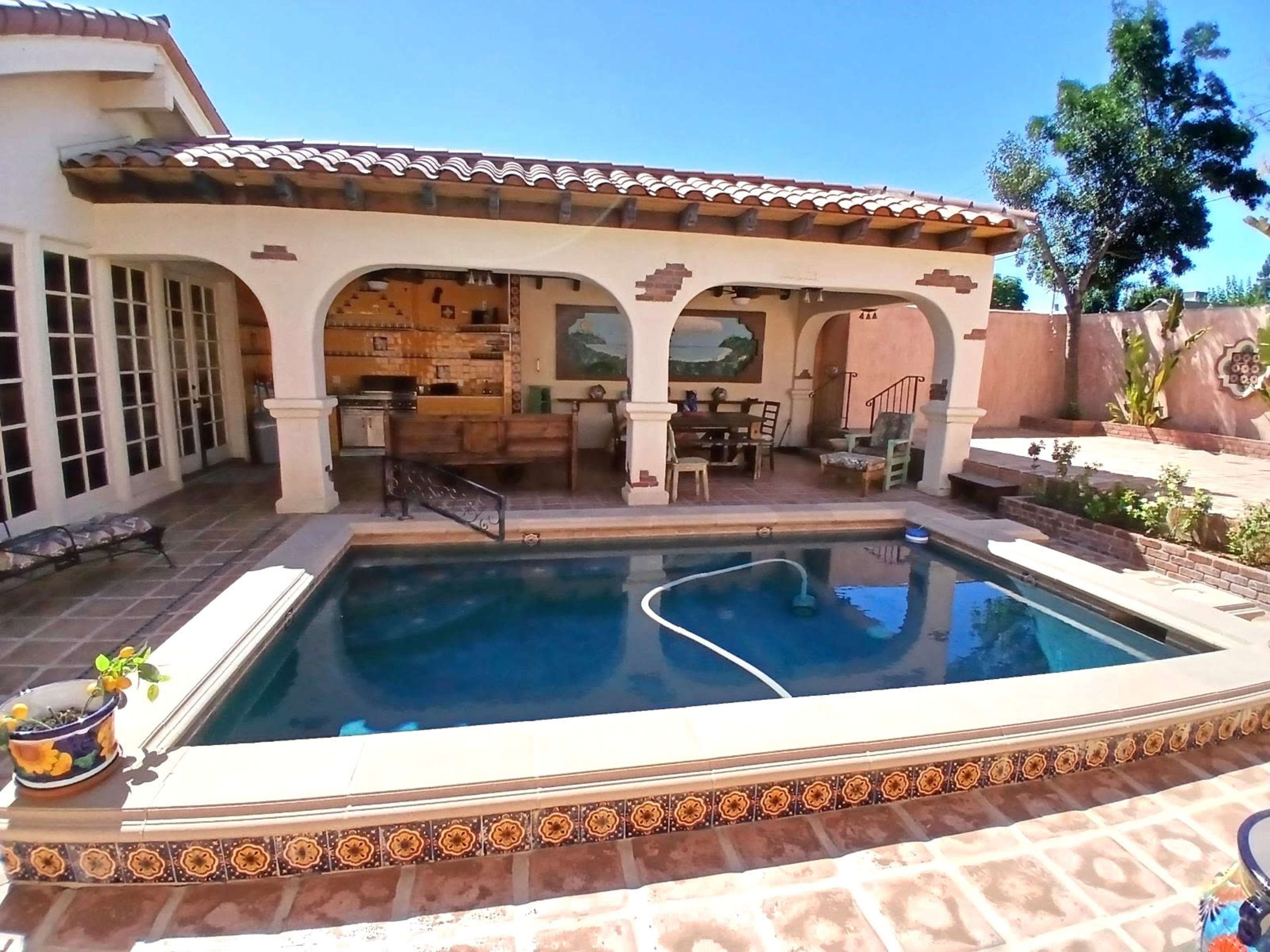 A courtyard with a pool, surrounded by a tiled patio and a covered outdoor seating area that includes a kitchen and dining space.