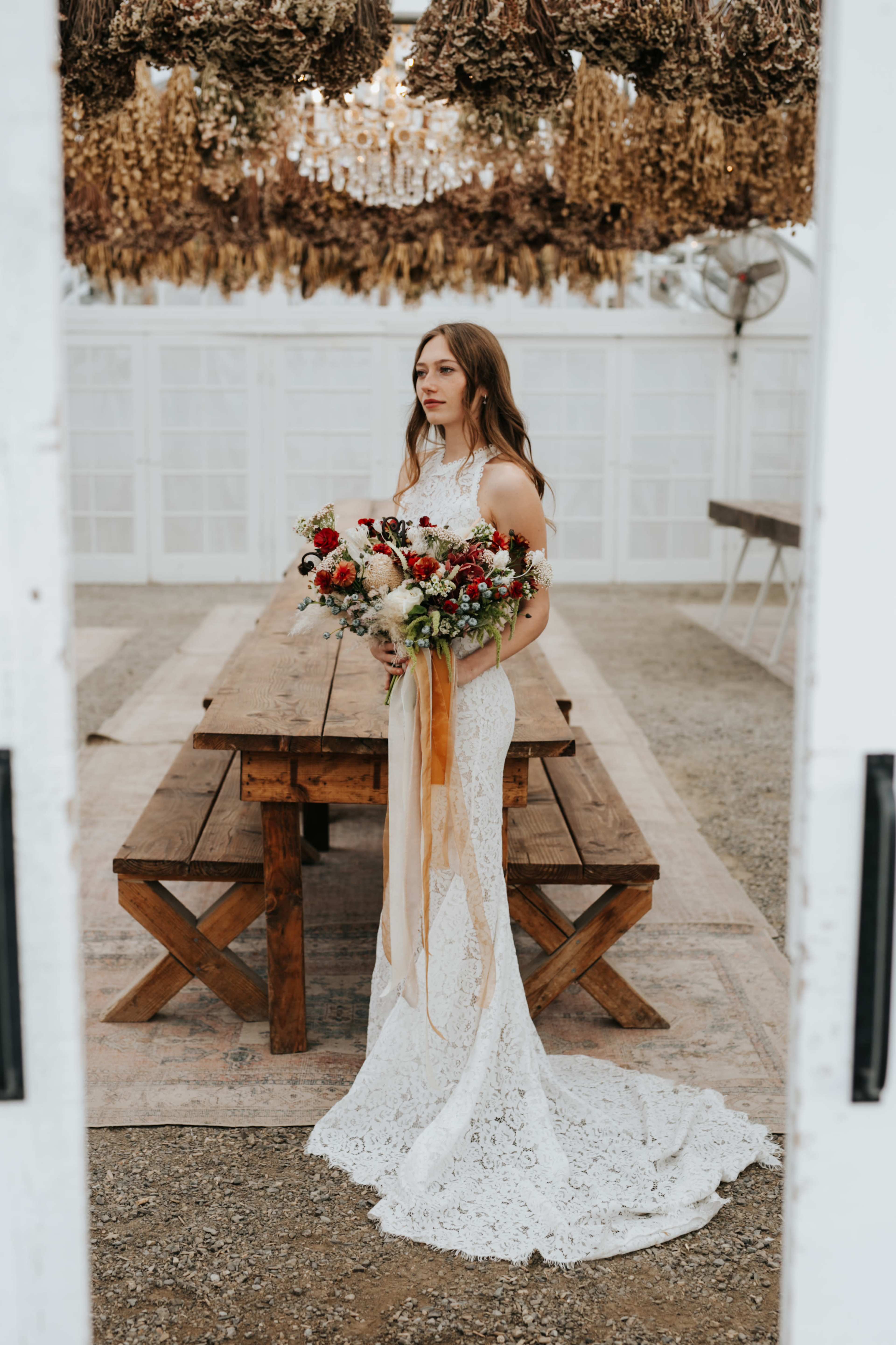 A bride in a lace wedding gown stands in a greenhouse holding a bouquet of flowers with long ribbons, surrounded by wooden tables and hanging dried plants.