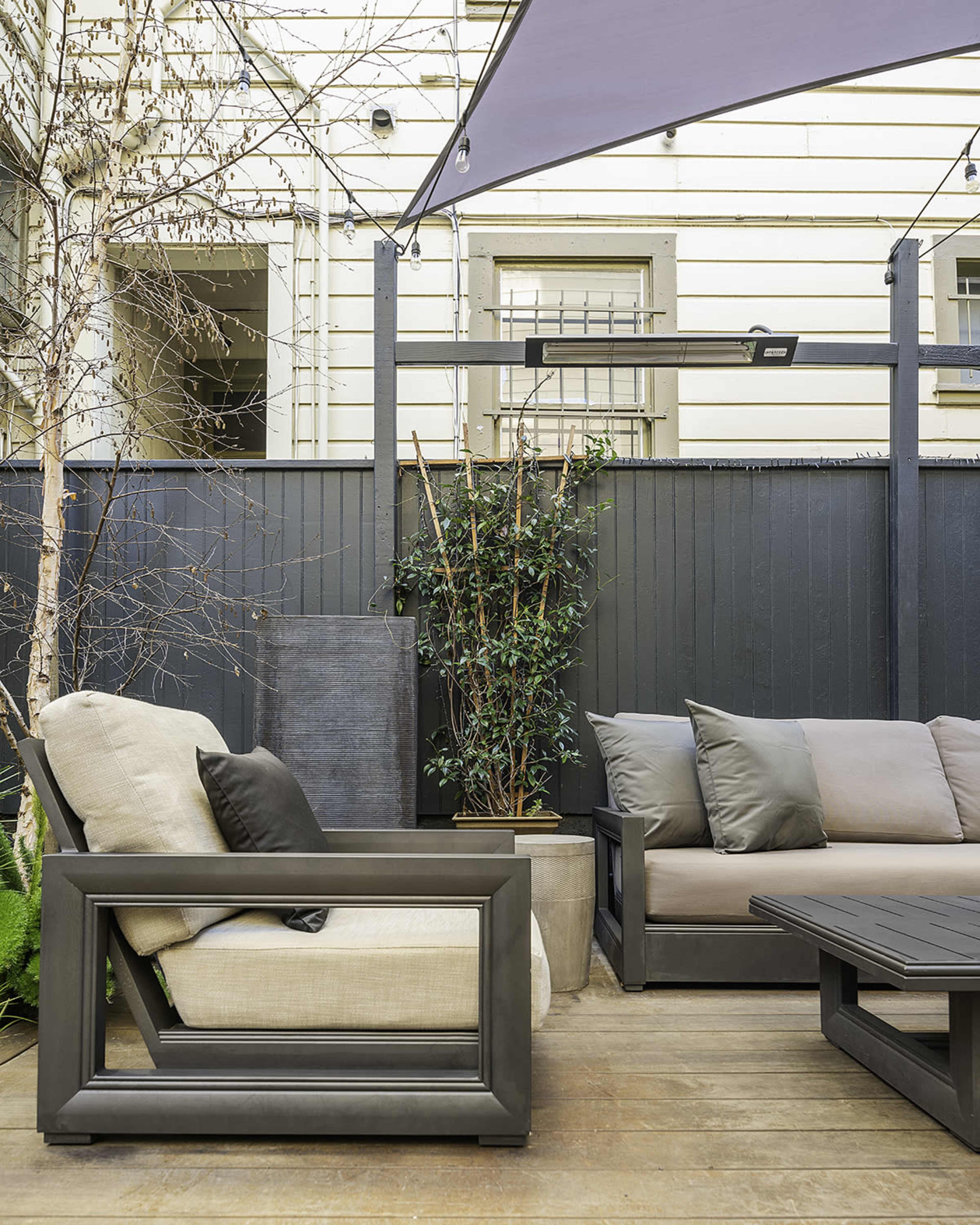 A small outdoor seating area with modern furniture, including a light-colored armchair and a dark sectional sofa, surrounded by a wooden deck and garden plants.