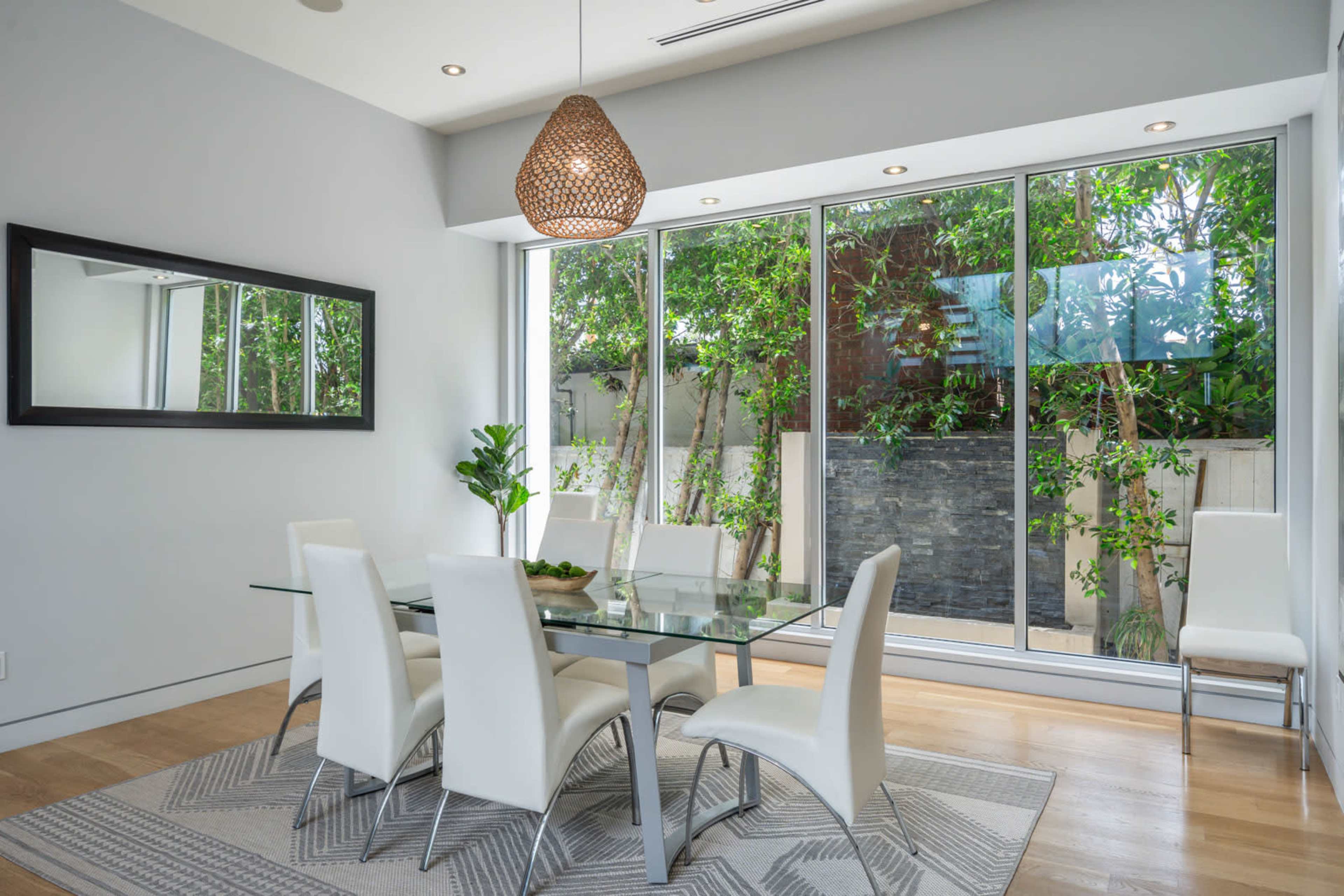 A dining area features a glass table surrounded by white chairs, with a large window showcasing greenery outside.