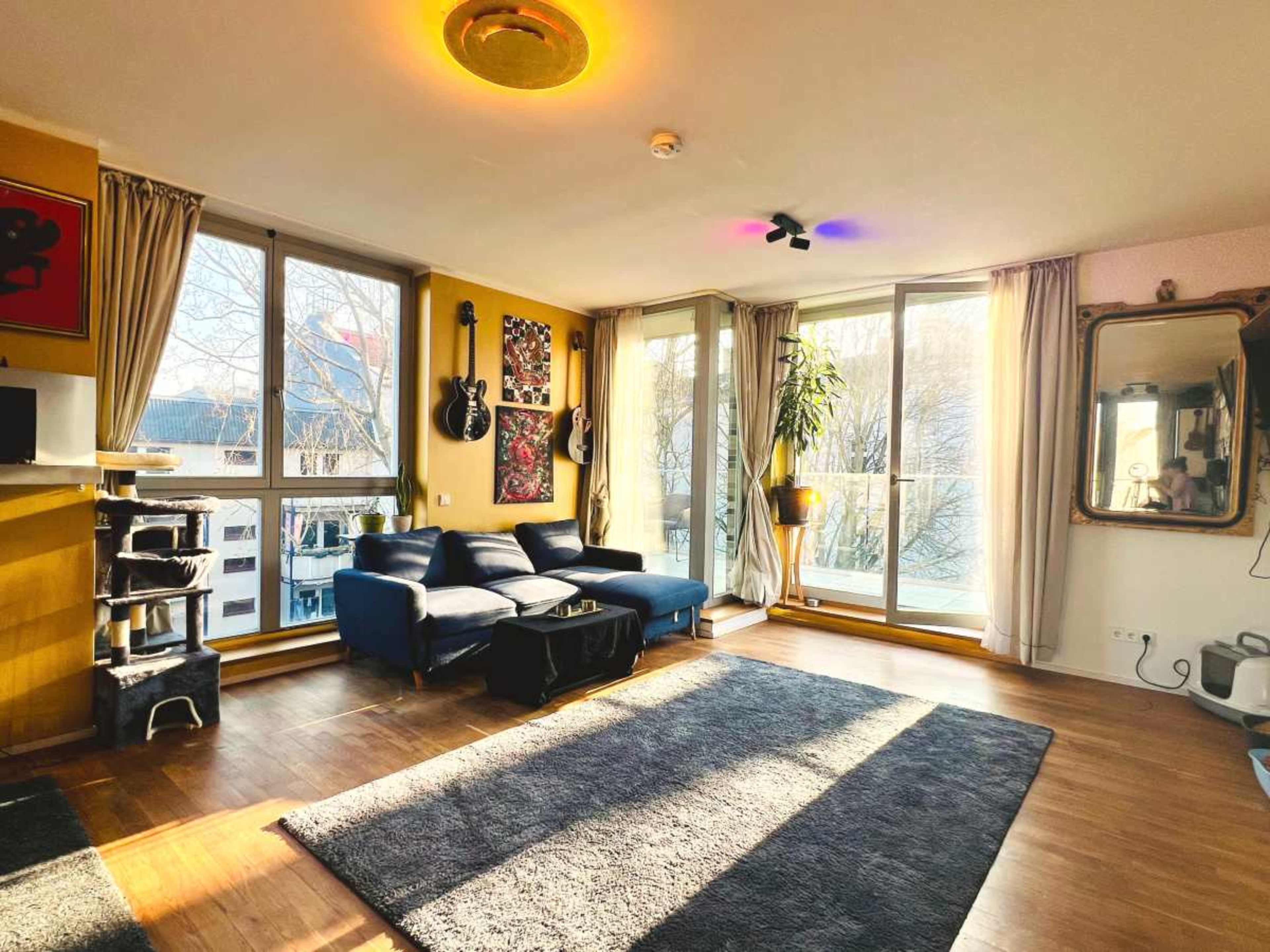 The image shows a bright living room with a sofa, large windows, a rug on the wooden floor, and decorative items on the walls.