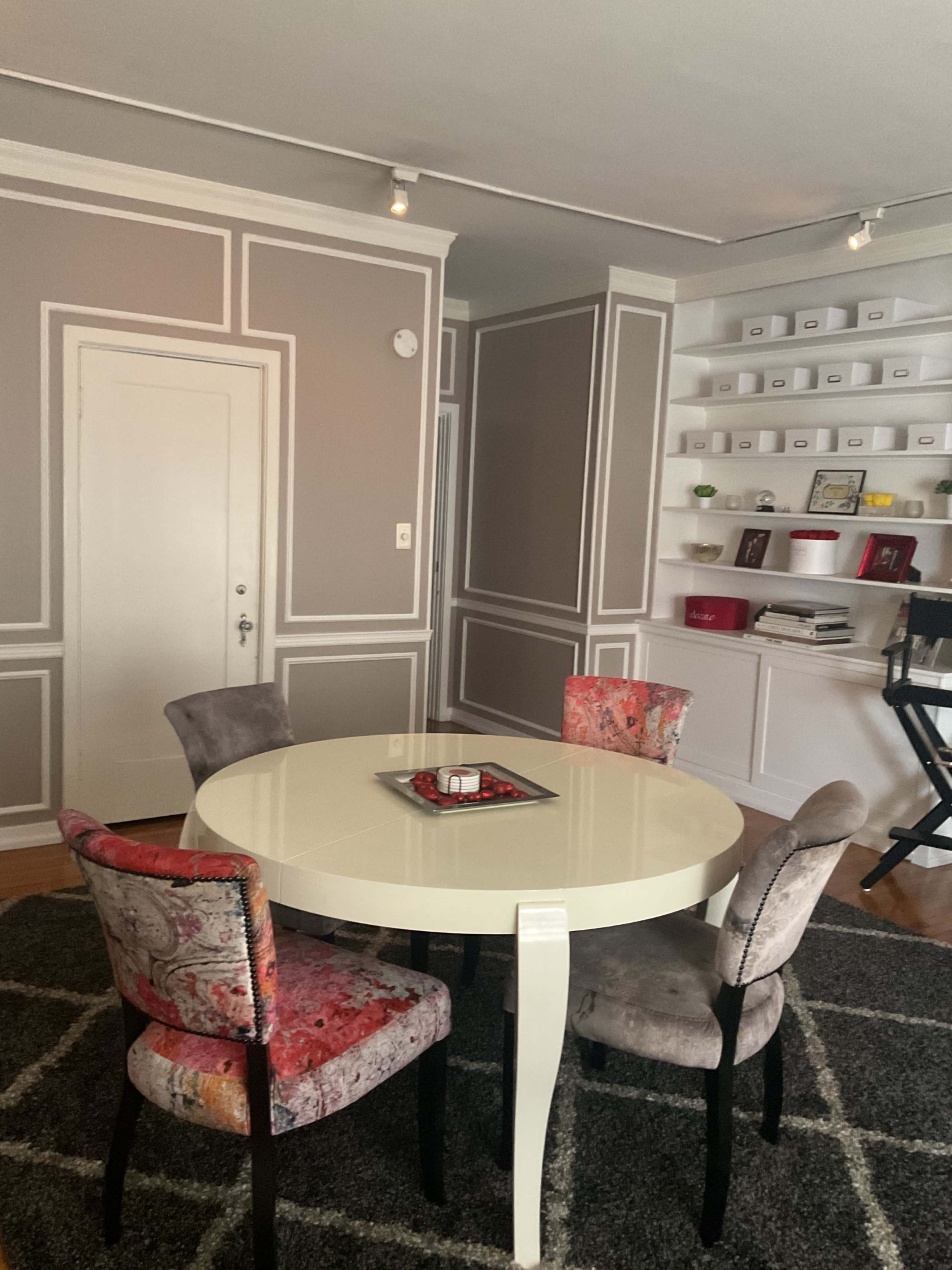The image shows a modern dining area with a round white table surrounded by four colorful chairs, against a backdrop of gray walls with white moldings and a bookshelf.