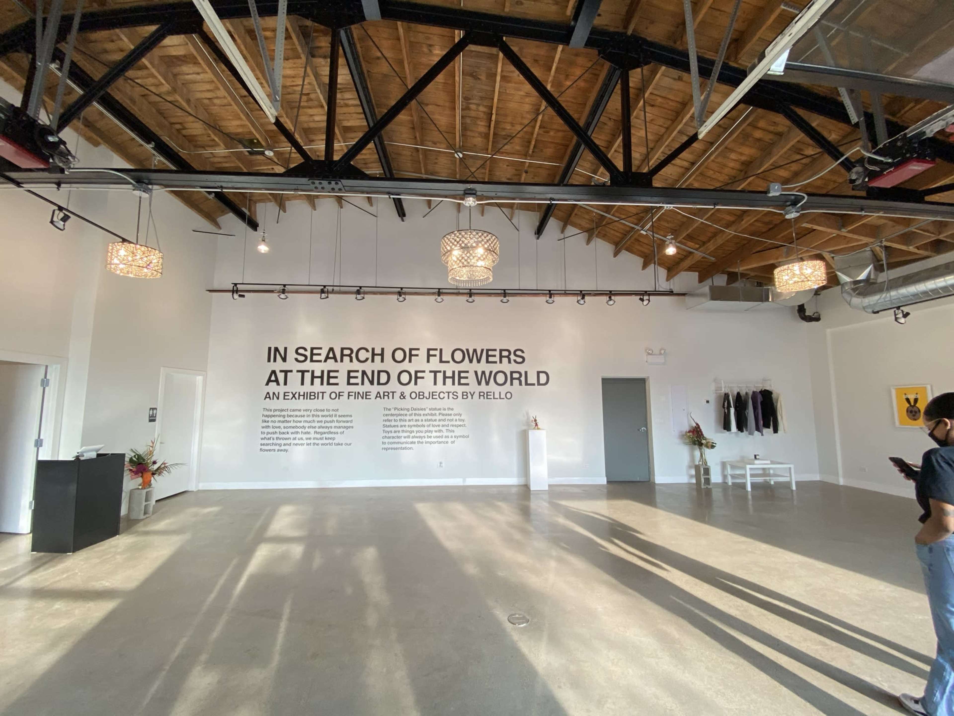 The interior of a gallery features a large wall with text announcing an exhibit titled "In Search of Flowers at the End of the World," along with a minimalist setup, a few decorative items, and a person standing to the side.
