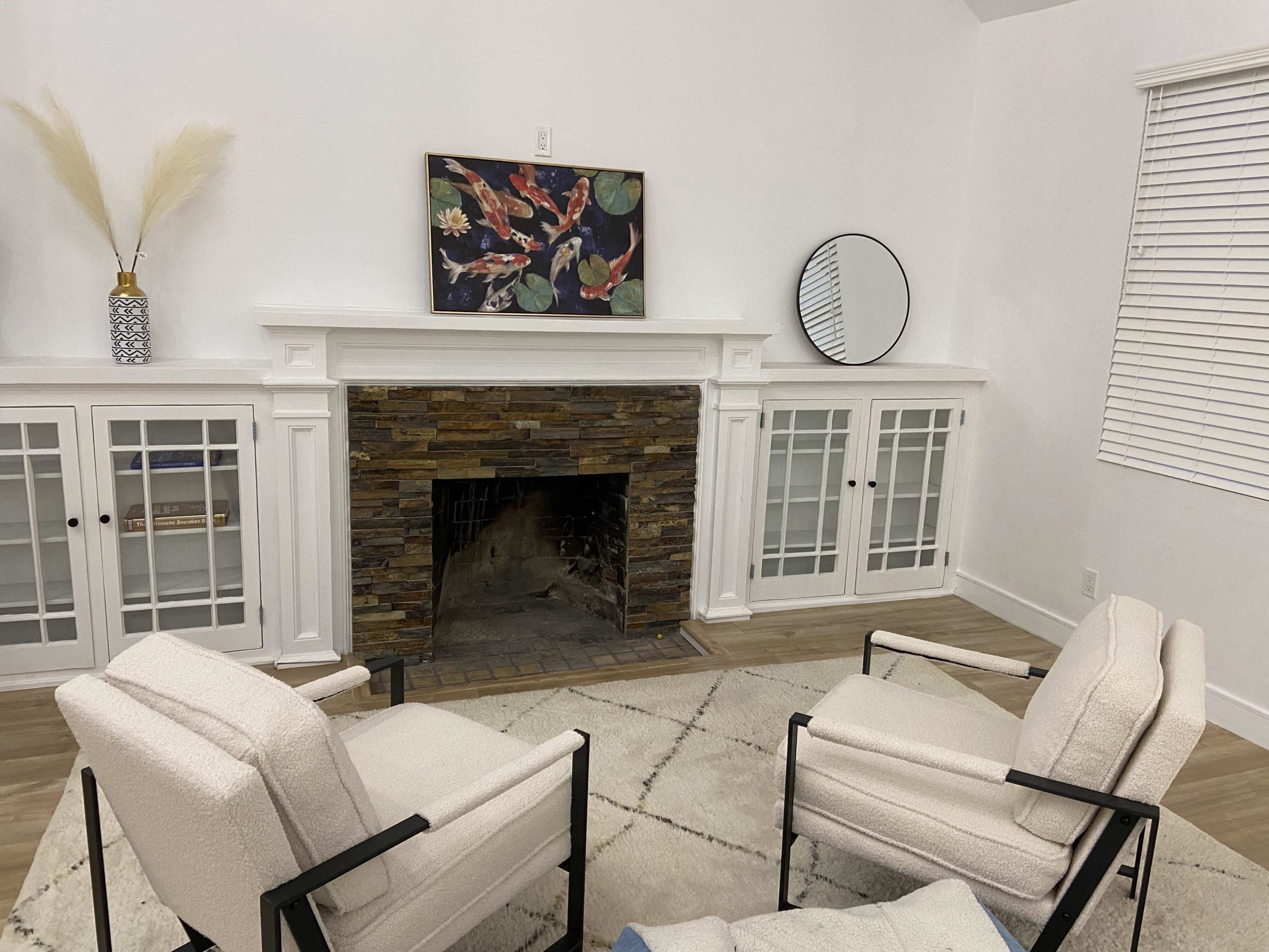 A living room features two chairs facing a stone fireplace, with a large decorative mirror and framed artwork on the wall above.