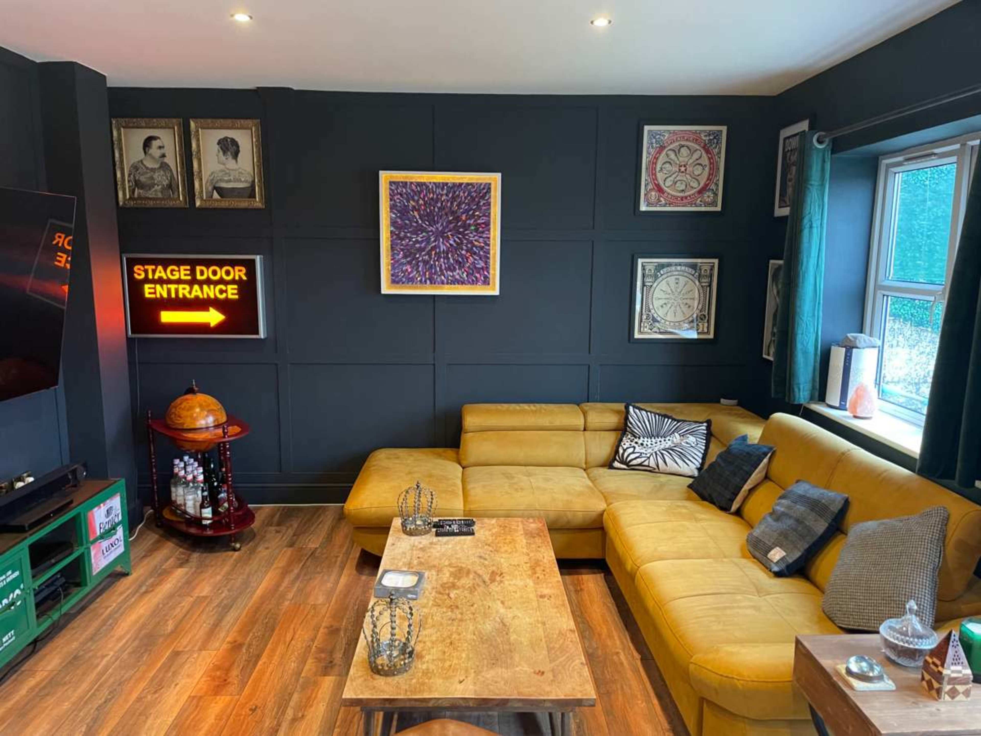 A cozy living room features a yellow sectional sofa, a wooden coffee table, and a wall decorated with framed artwork and a neon sign that reads "STAGE DOOR ENTRANCE."