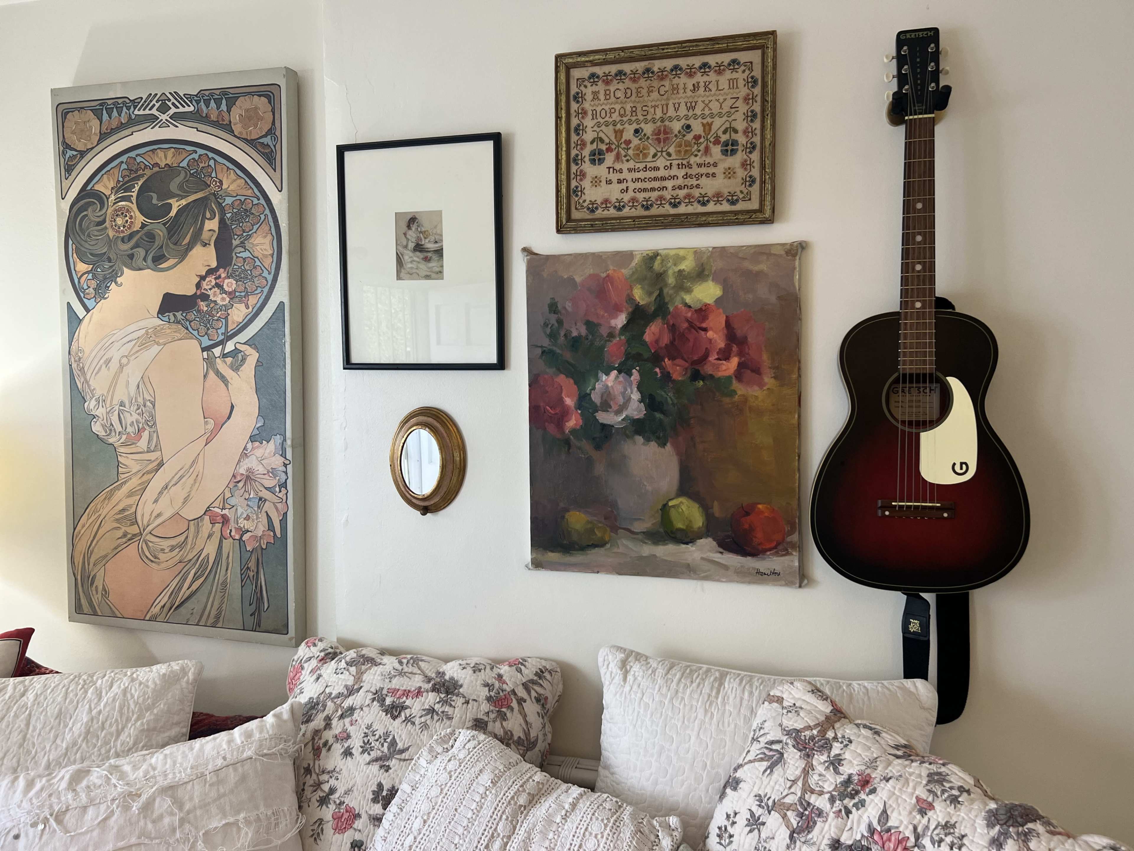 A wall decorated with various pieces of art, including a vintage poster, a framed photograph, a floral painting, and a guitar hanging beside a small mirror.