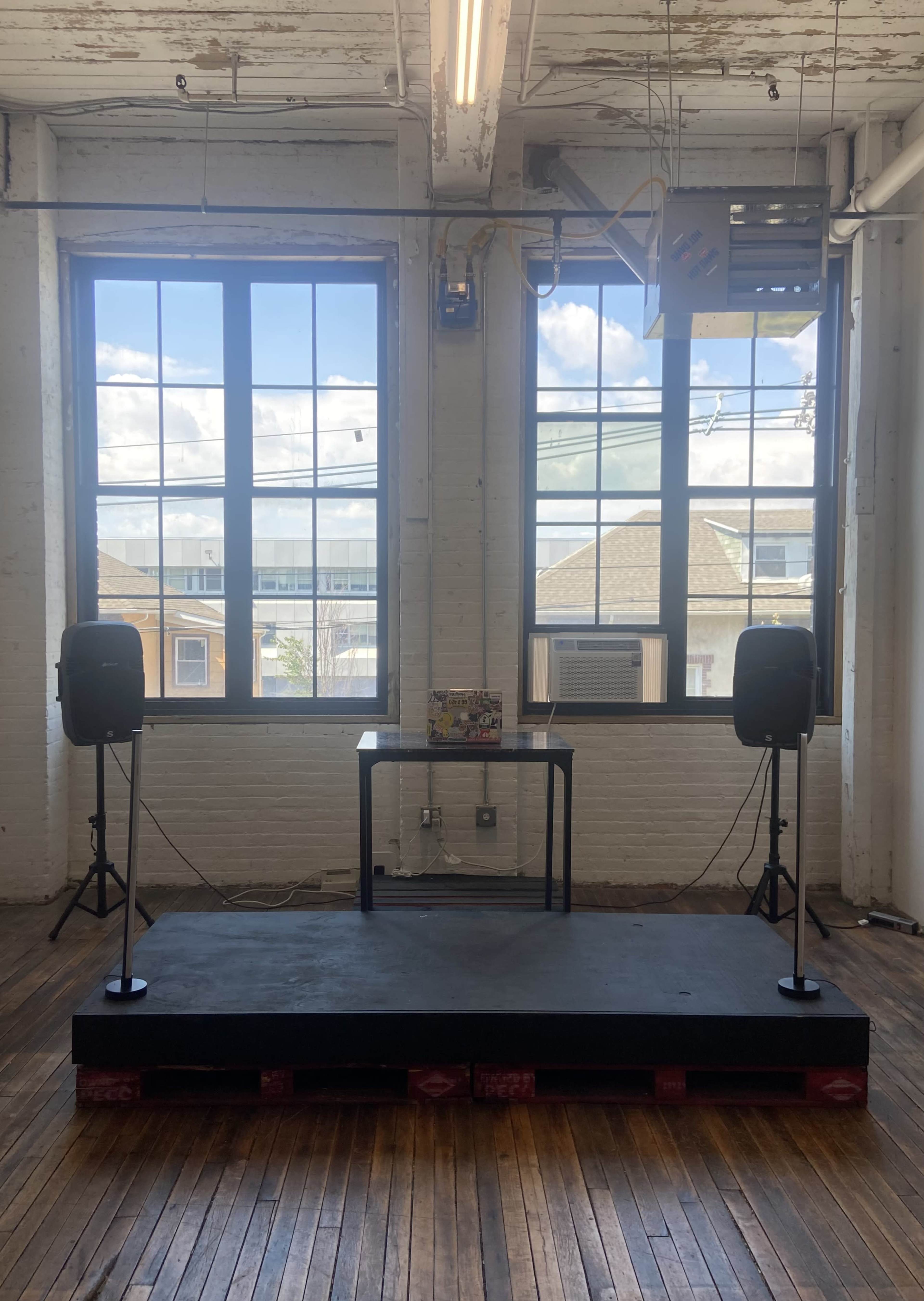 The image shows a small stage made of pallets with two speakers on either side, set in a room with large windows that overlook a neighborhood.