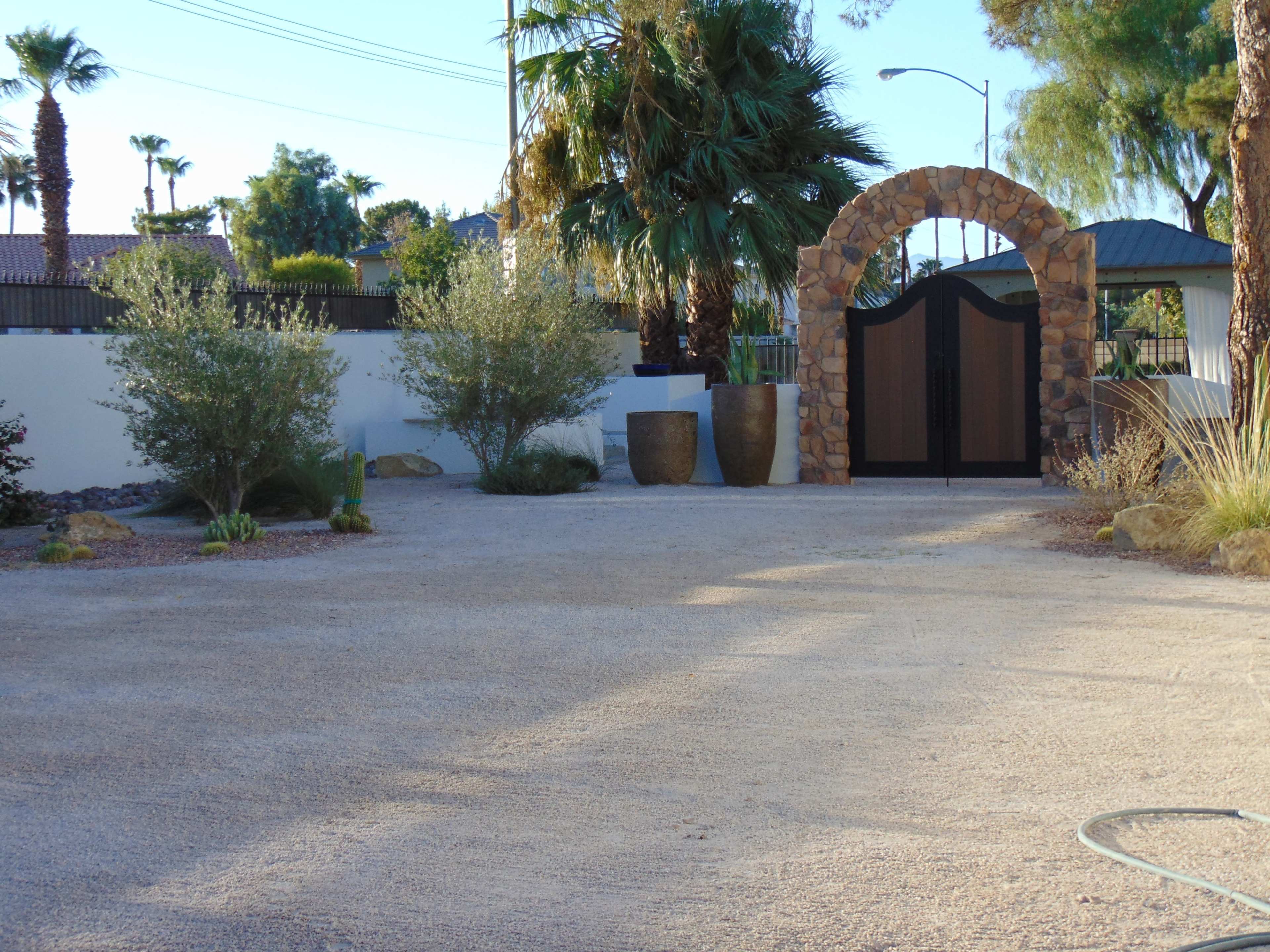 The image shows a gravel driveway leading to a stone archway with a gated entrance, flanked by potted plants and palm trees.