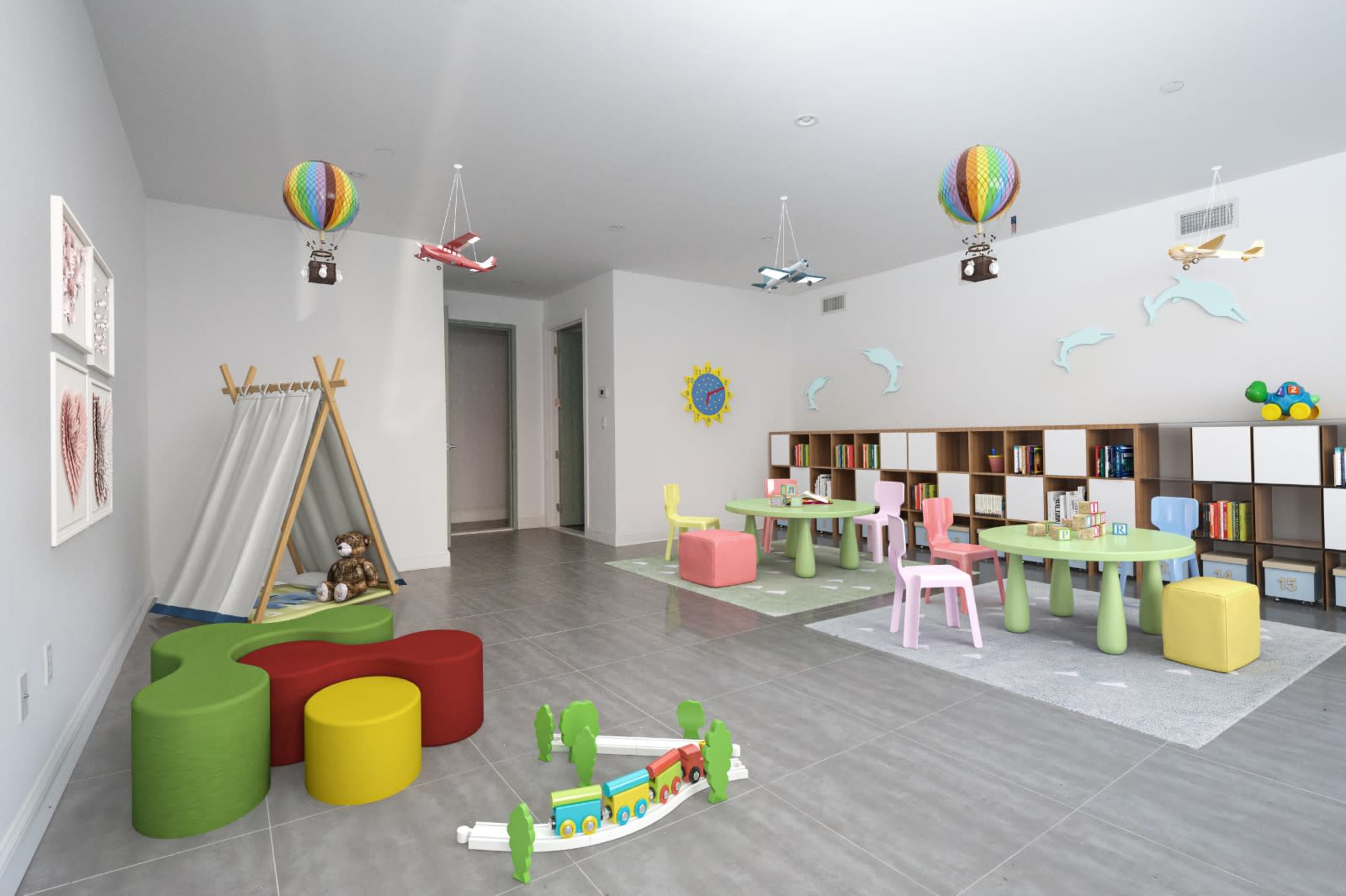 A colorful playroom featuring a teepee, various toy tables and chairs, floating decorative hot air balloons, and shelves filled with books and toys.