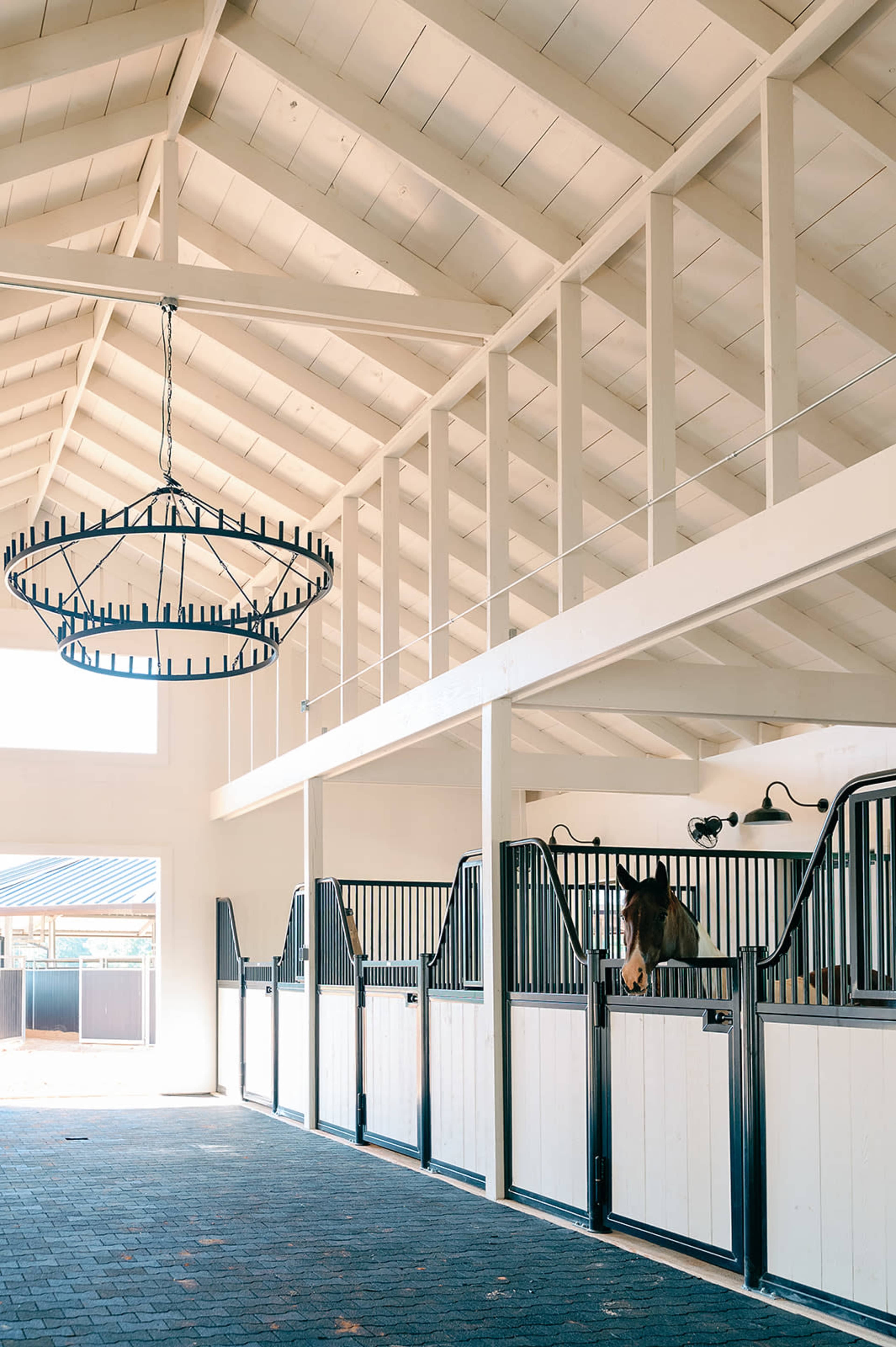 The image shows the interior of a horse stable with several stalls separated by black gates and a large chandelier hanging from a high ceiling.