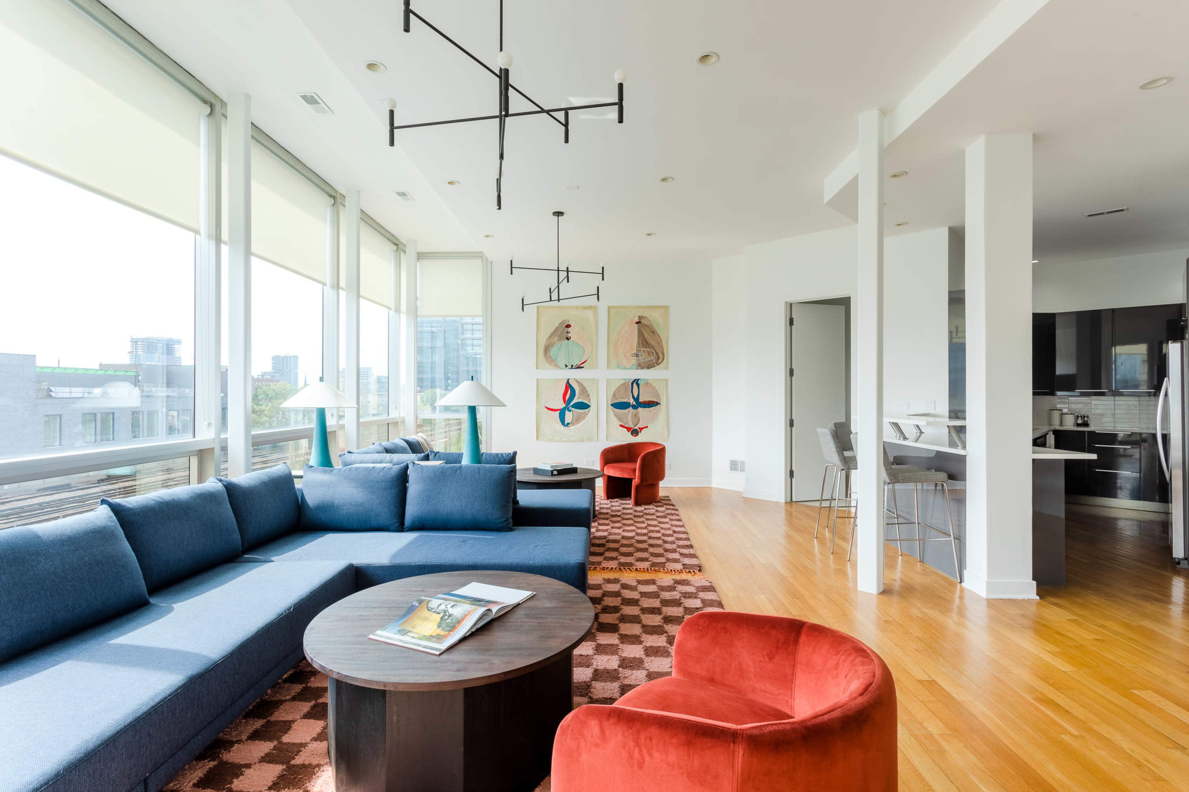 A modern living room with large windows, a blue sectional sofa, a round coffee table, and two red armchairs, featuring abstract art on the walls.
