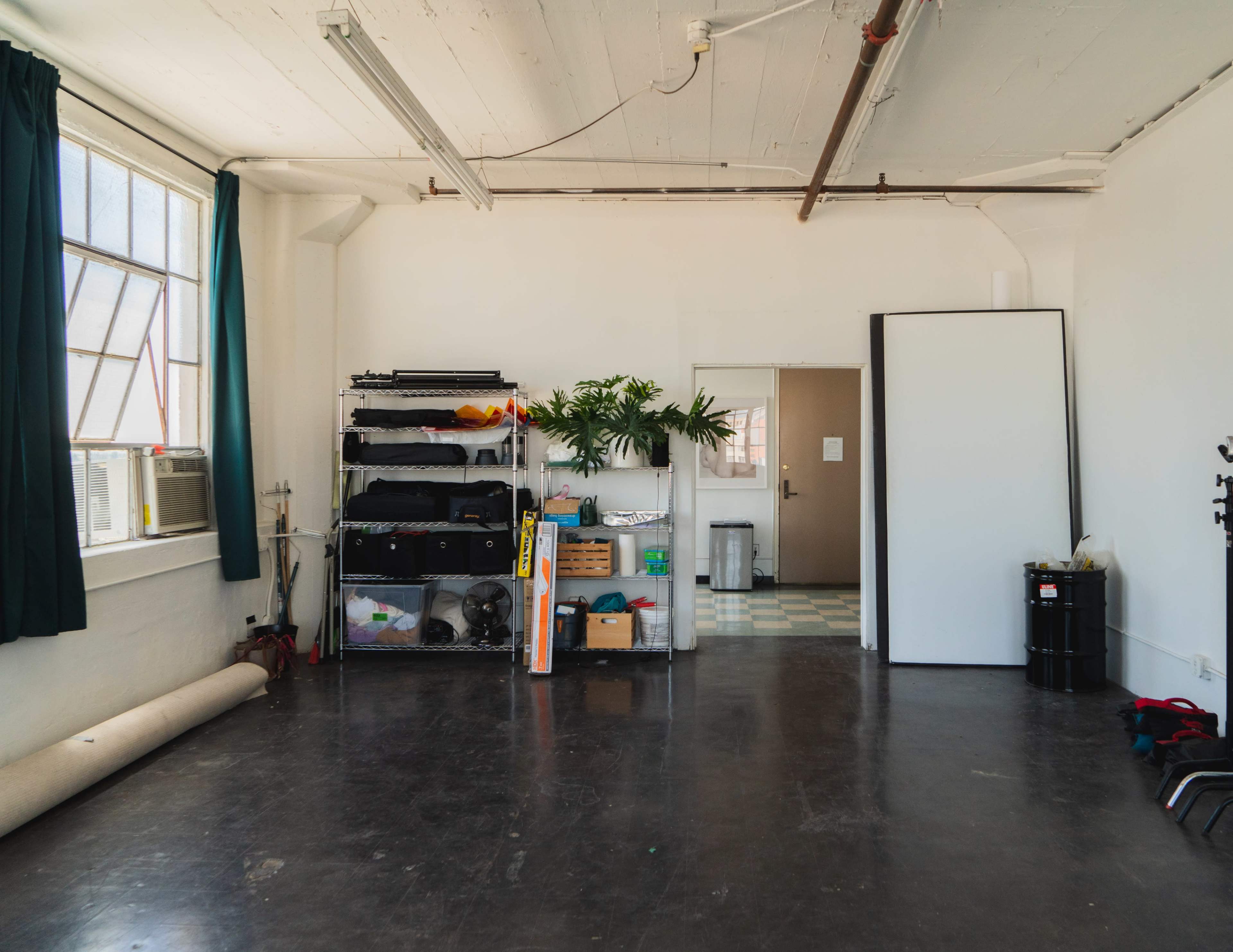The image shows an empty industrial-style room with a window, shelving units filled with items, and a door leading to another area.