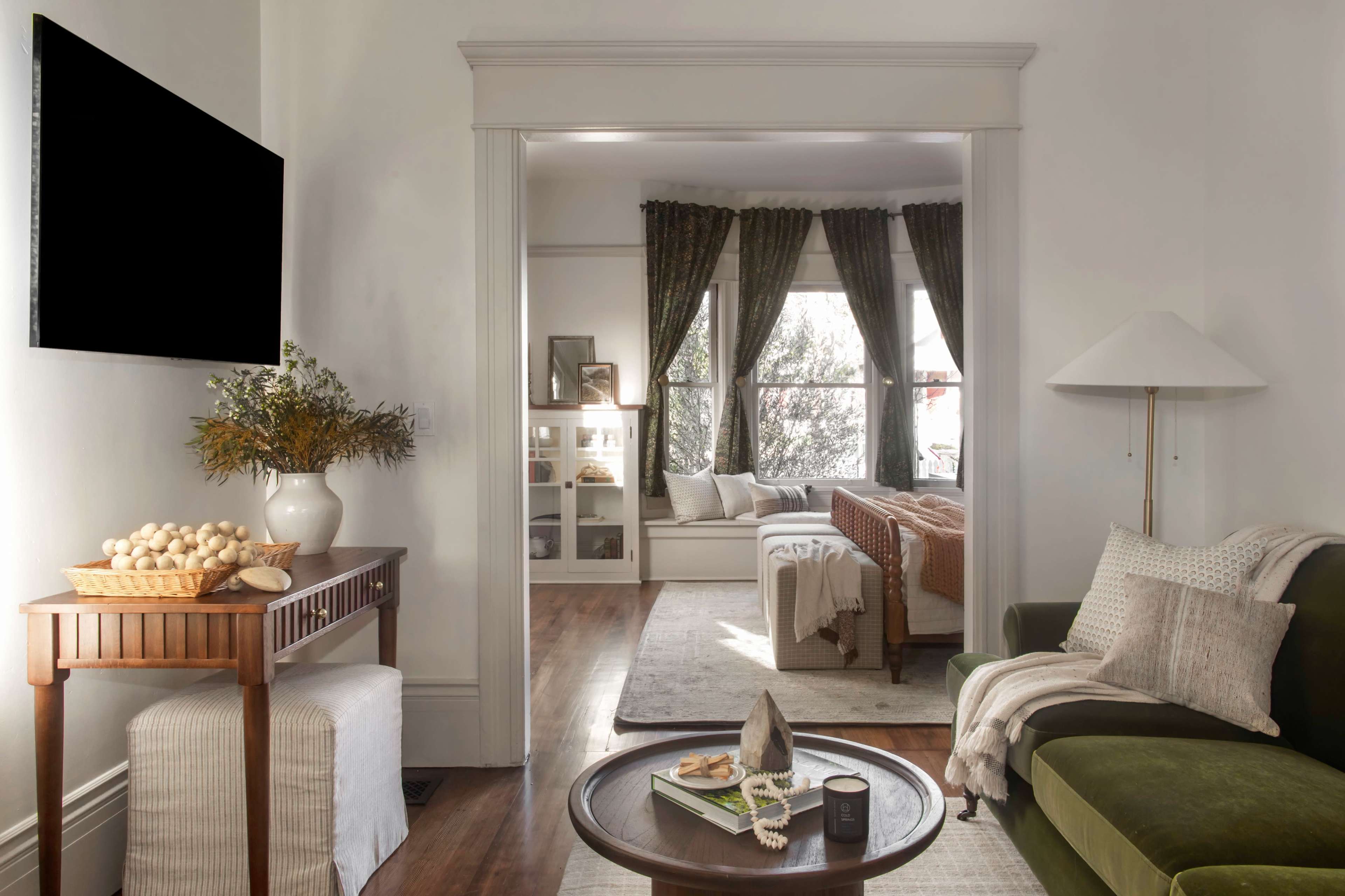 A living space features a green sofa, a wooden table with decorative items, and a view into a well-lit room with large windows and a bed in the background.
