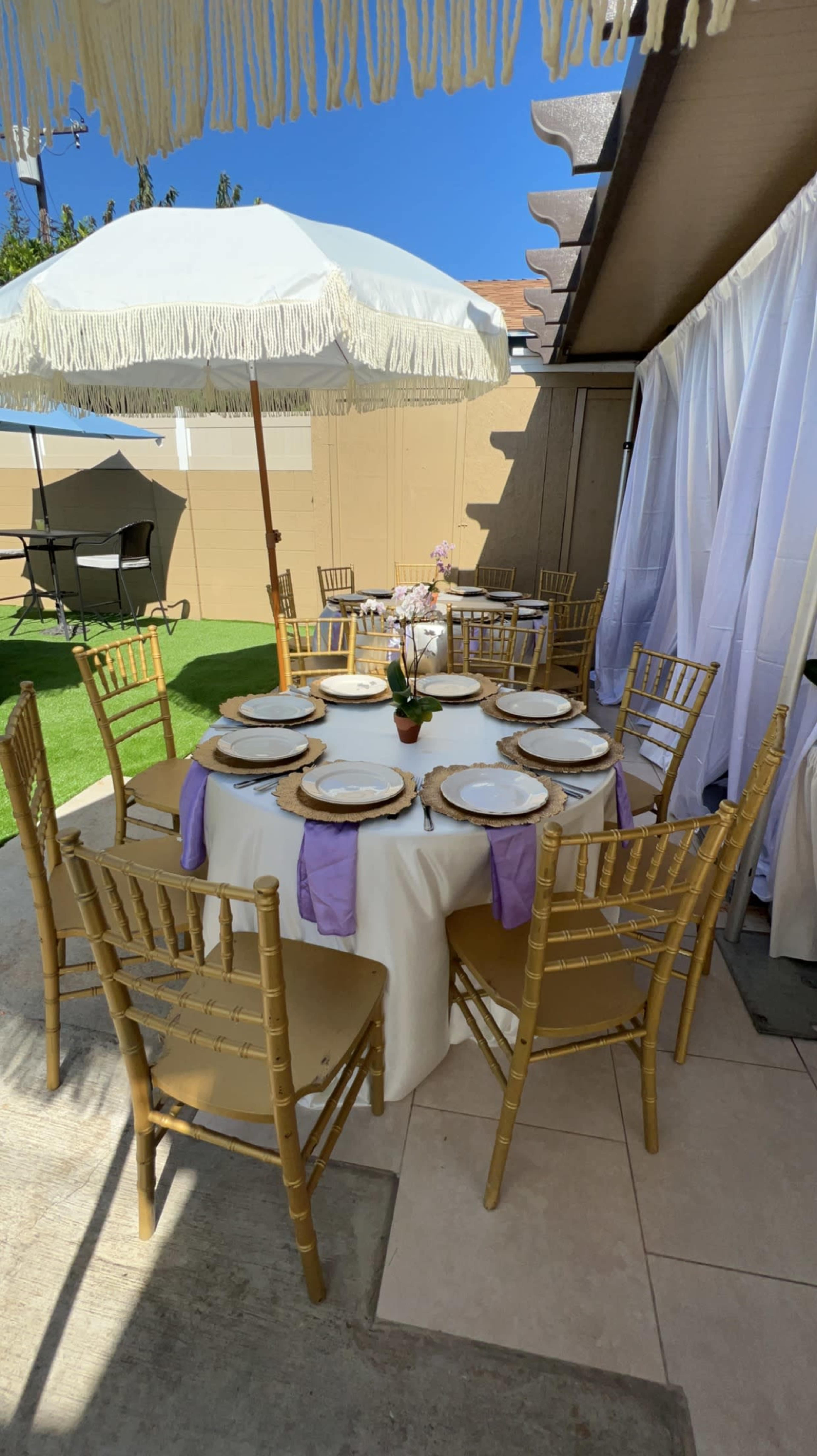 A round table set for a gathering is adorned with plates and decorative centerpieces, surrounded by gold chairs under a white parasol.