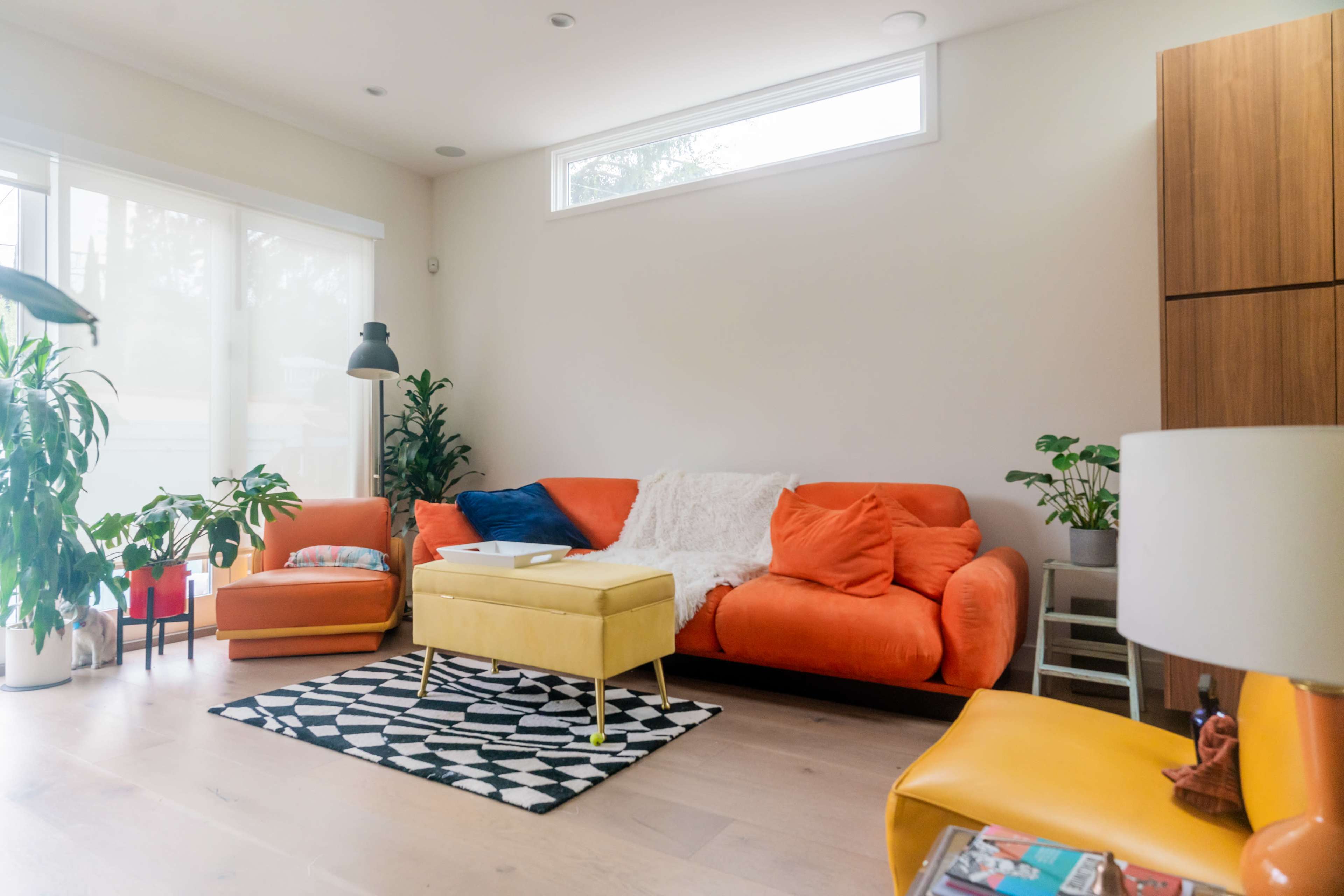 The image shows a modern living room featuring an orange sofa, a yellow ottoman, and several green plants arranged around a geometric-patterned rug.