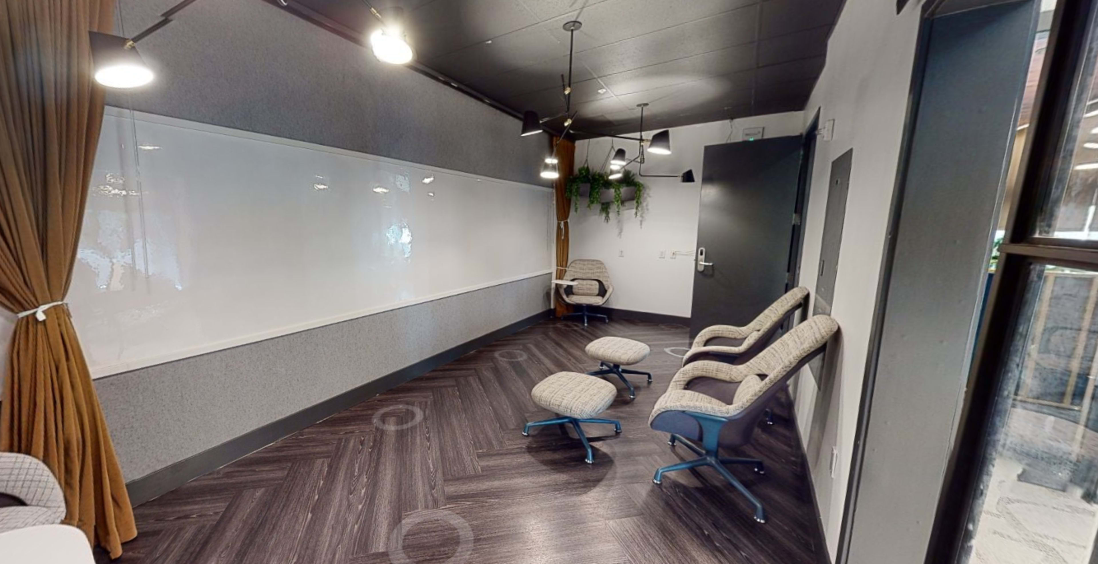 A small meeting room features a whiteboard, four wheeled chairs arranged in a semicircle, and industrial-style lighting.