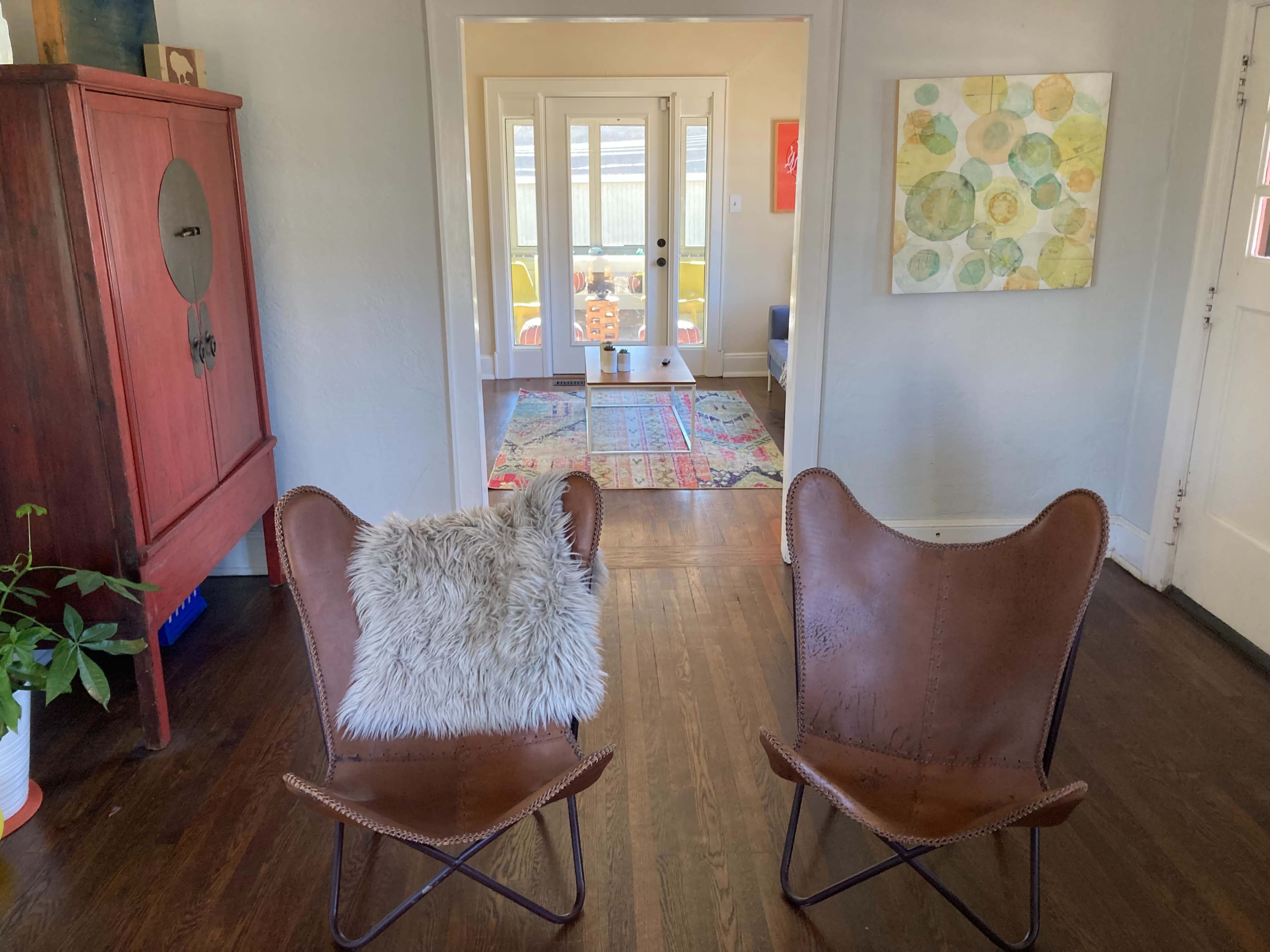 Two leather butterfly chairs with a throw pillow face a hallway leading to a brightly lit room with a colorful rug and doorway.
