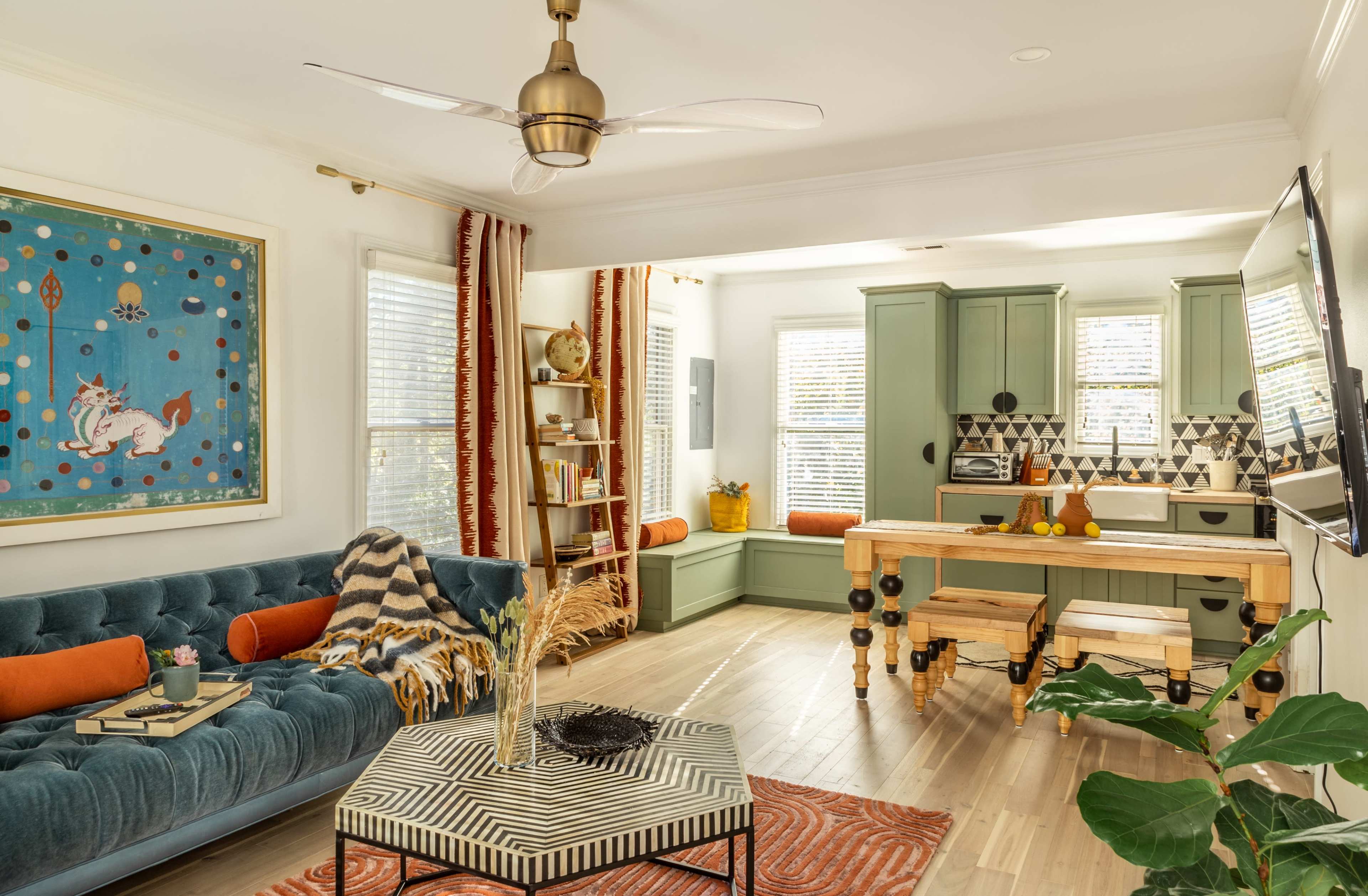 A contemporary living space featuring a blue velvet sofa, an adjoining kitchen with green cabinetry, and large windows that allow natural light to fill the room.