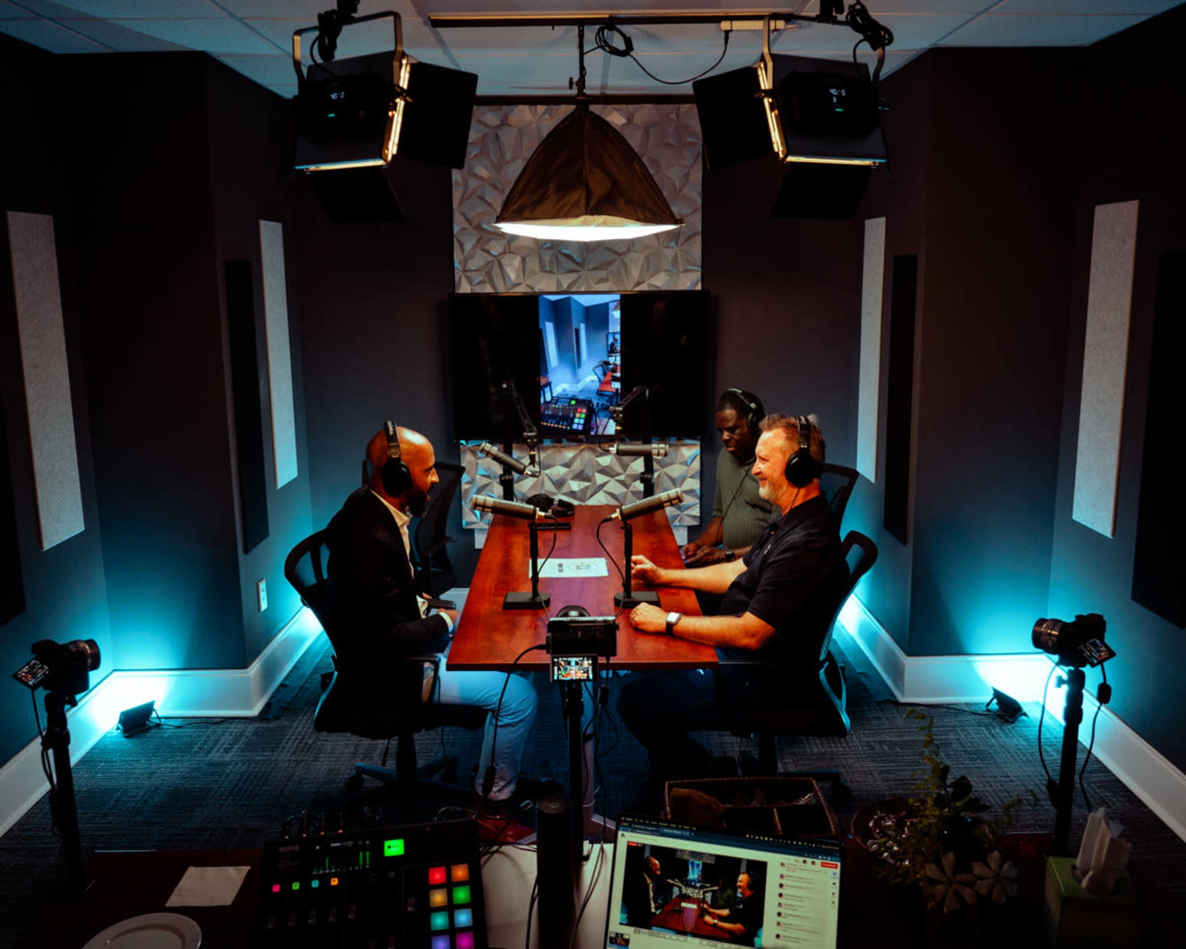 Three people sit around a wooden table in a modern podcast studio, equipped with microphones and camera equipment, while a screen in the background displays a digital interface.