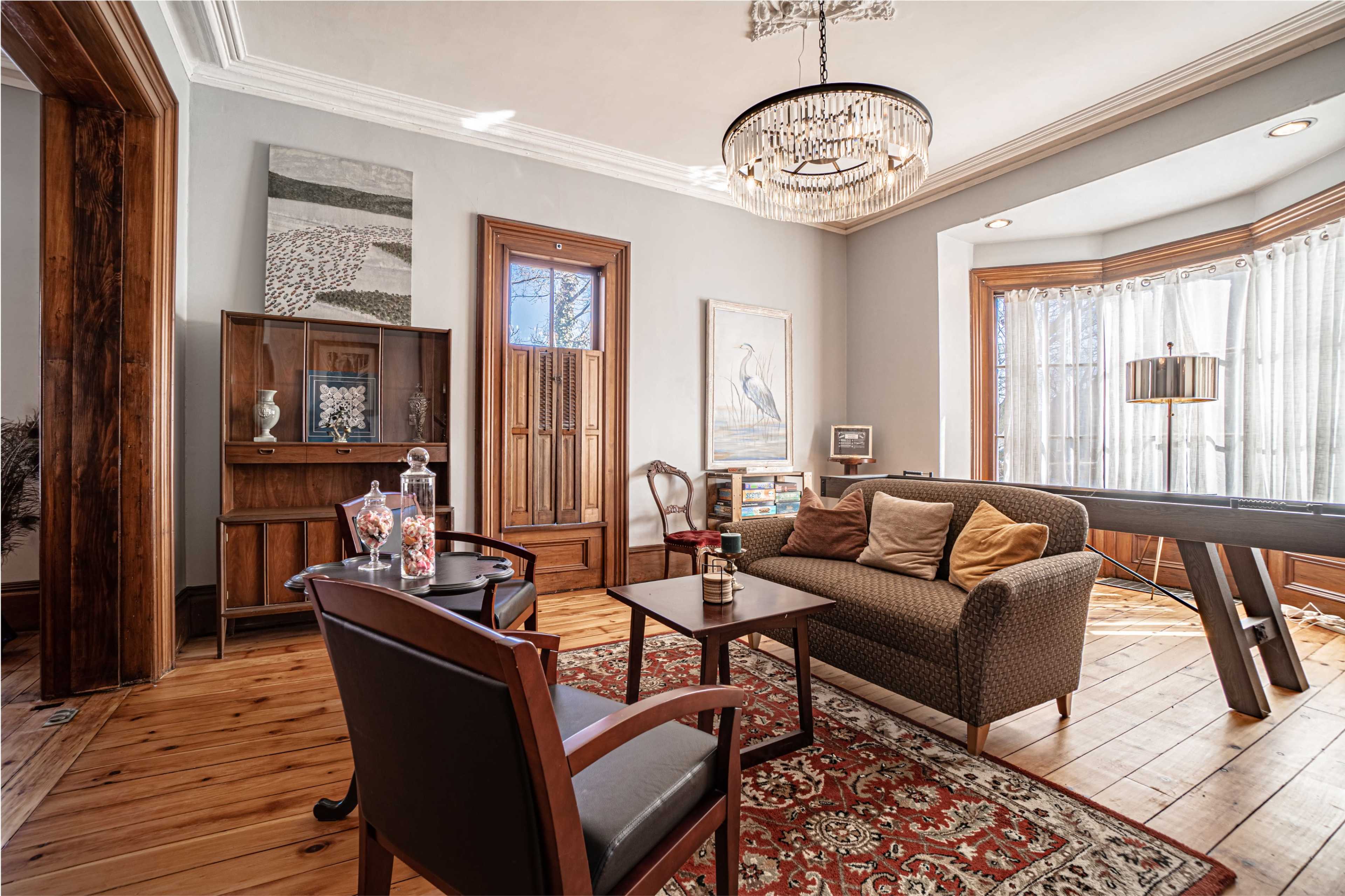 The living room features a sofa with cushions, a wooden cabinet, a chandelier, and large windows with sheer curtains.