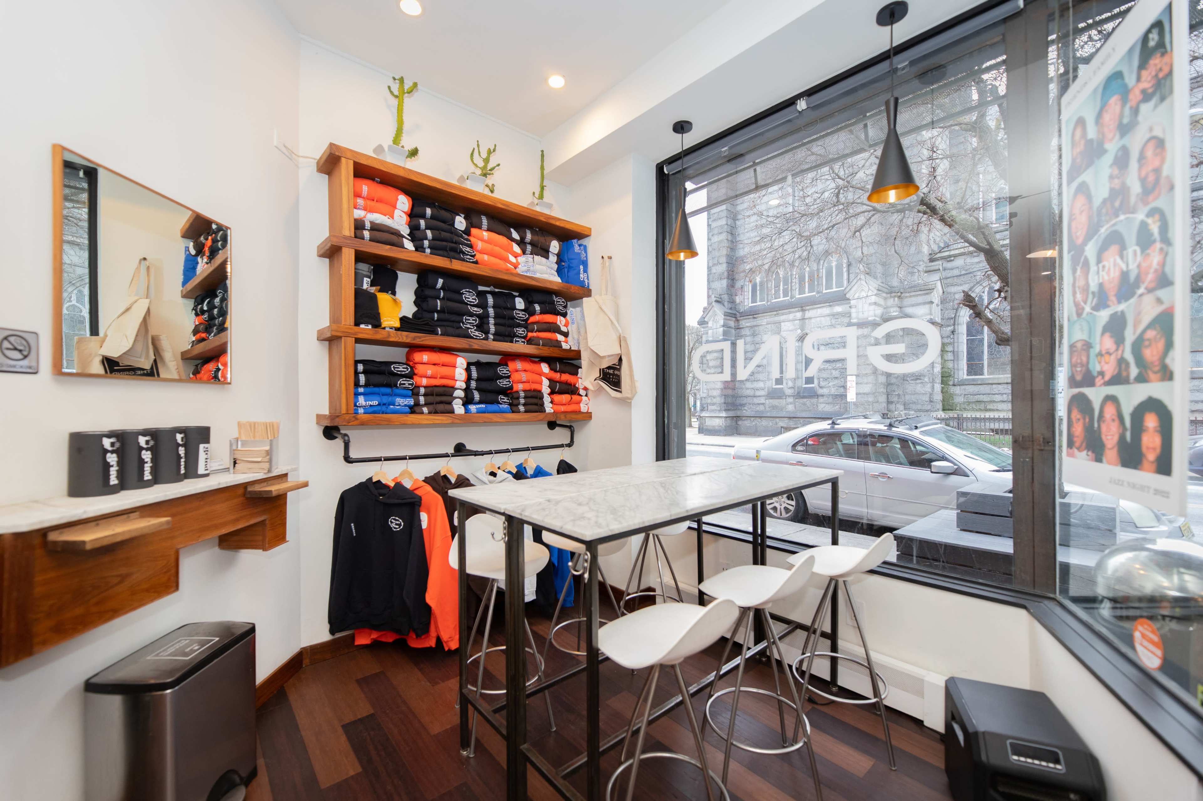 A clothing store features a display of stacked shirts in various colors on a wooden shelf, with a small seating area and large windows showing the street outside.