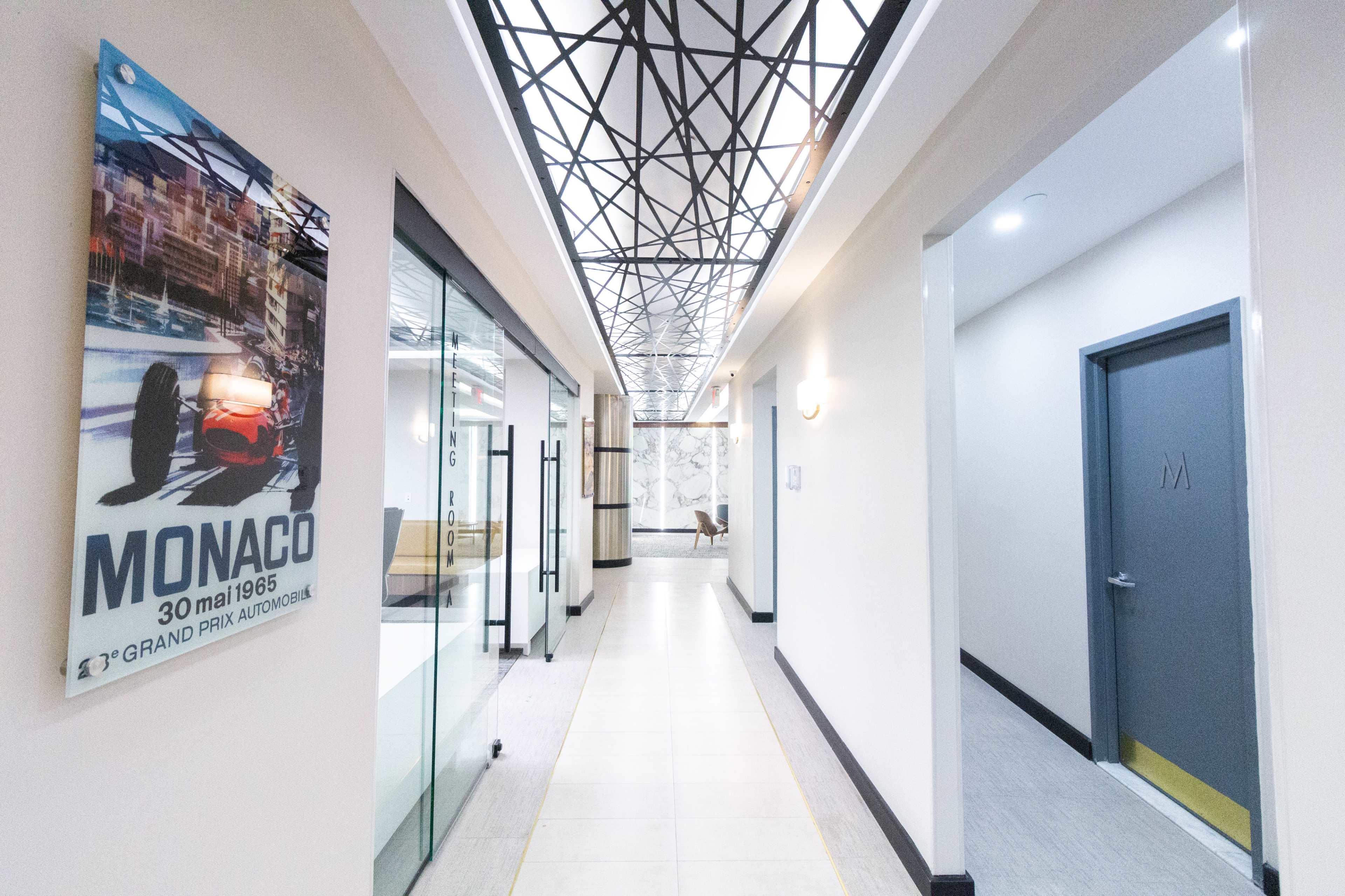 A well-lit corridor features glass-walled offices on one side and doorways to private rooms on the other, adorned with a decorative sign for the Monaco Grand Prix.