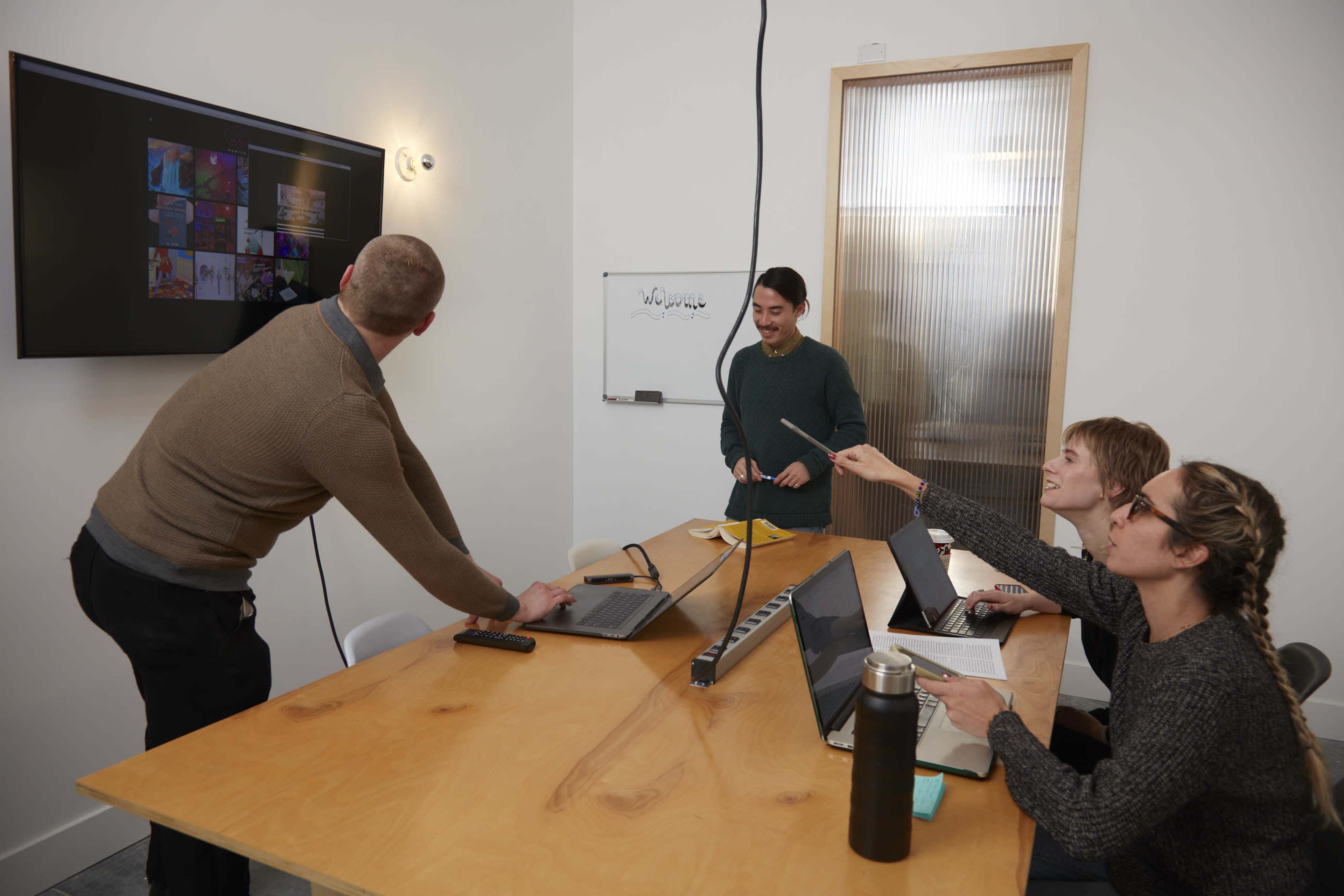 A group of four people collaborates at a meeting table, with one person presenting on a screen while others engage with laptops and notes.