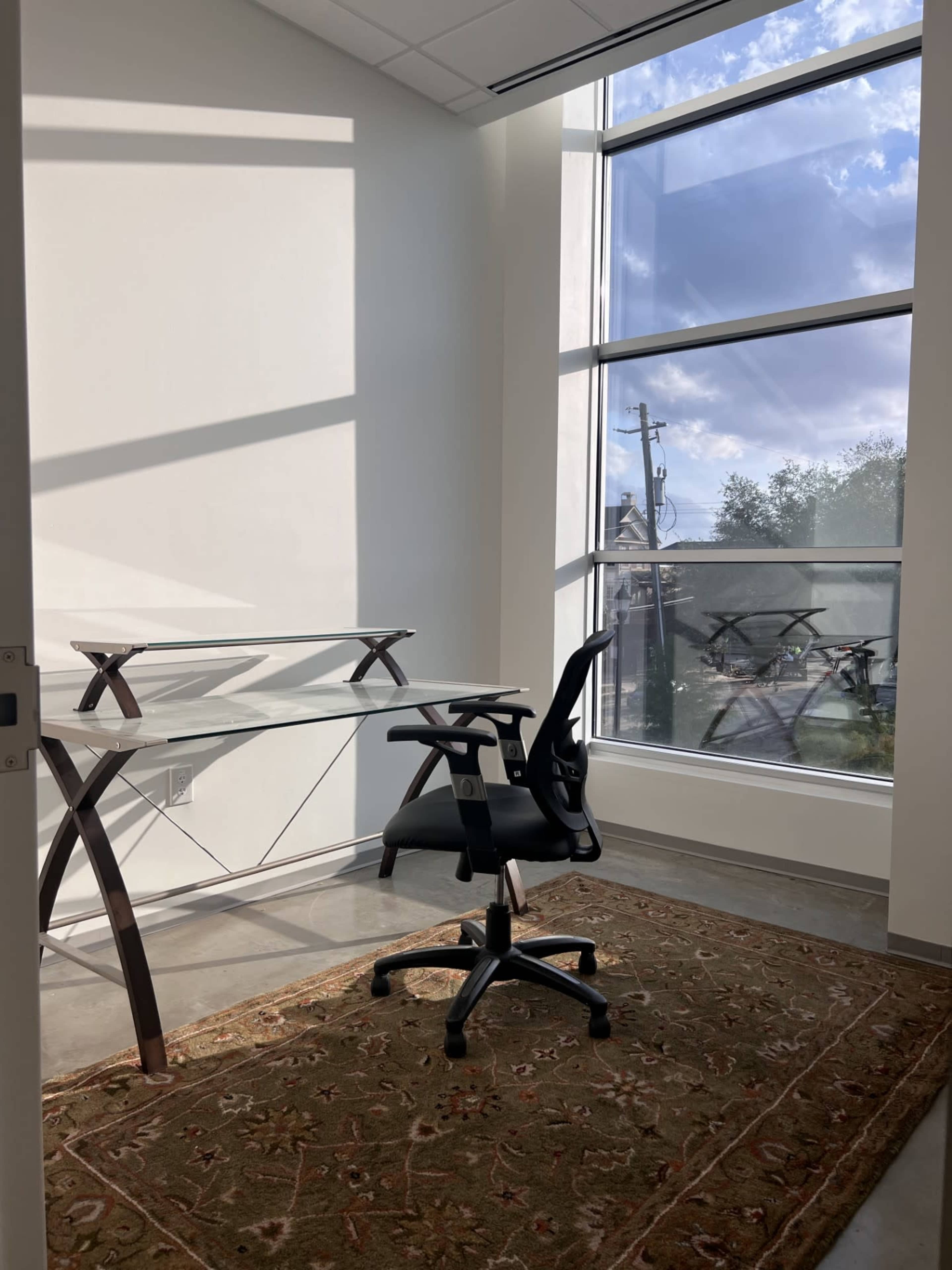 The image shows a small office space featuring a glass desk, a black office chair, and a large window with natural light and a view outside.