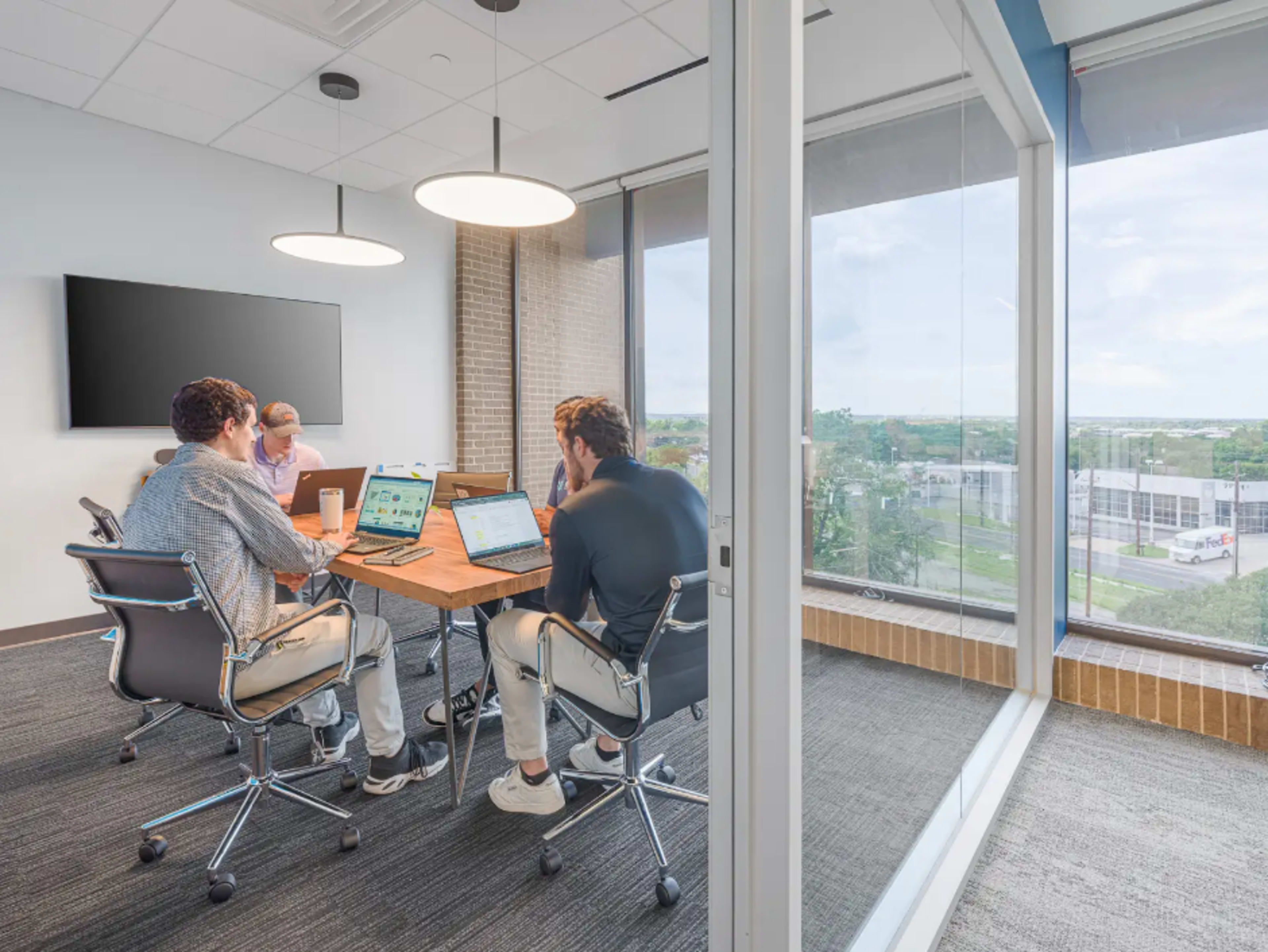Three professionals are seated around a conference table with laptops in a glass-walled meeting room, while another person works nearby.