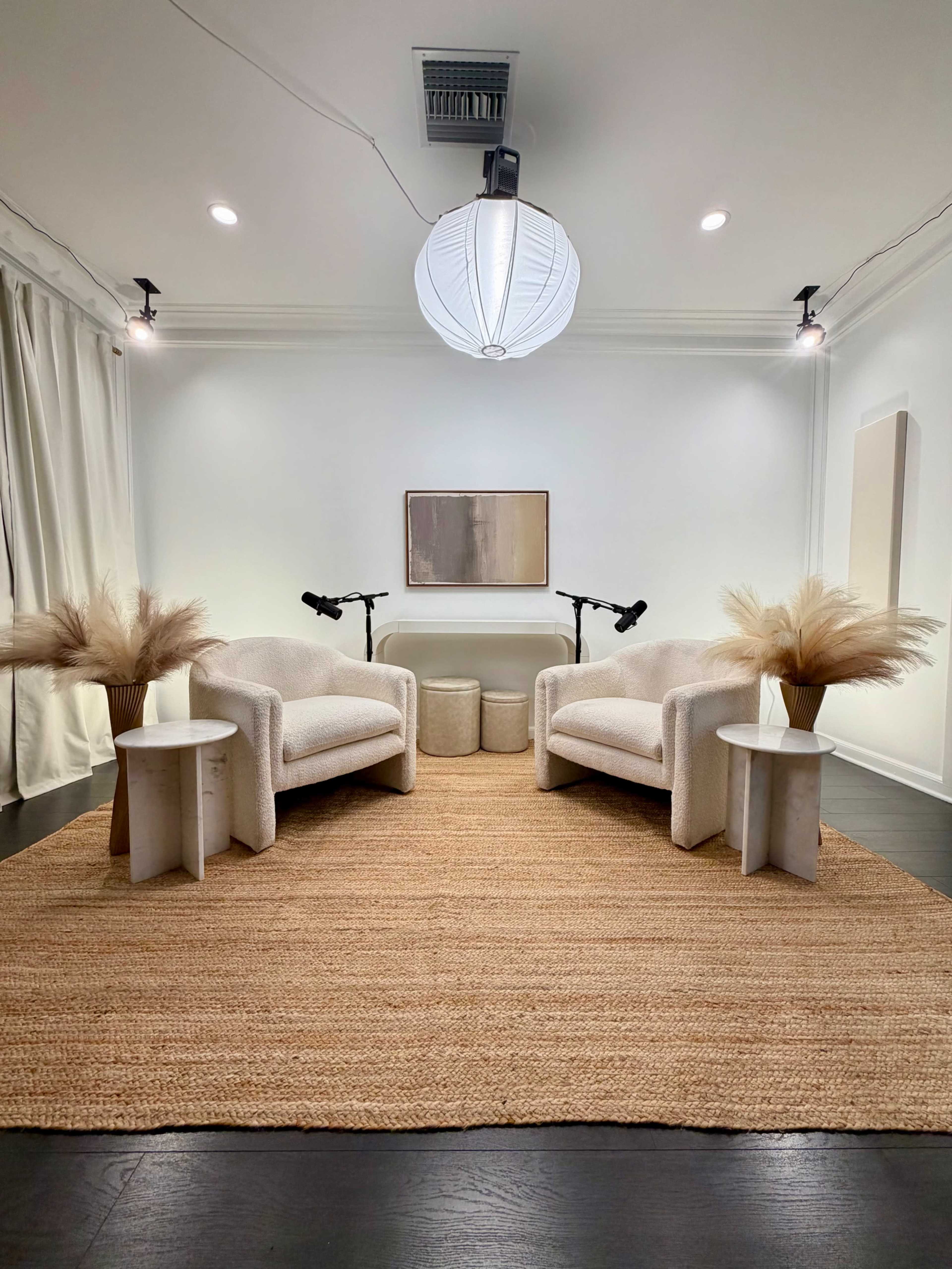 The image shows a minimalist living space with two upholstered chairs, side tables, and decorative pampas grass arranged on a natural fiber rug under overhead lighting.