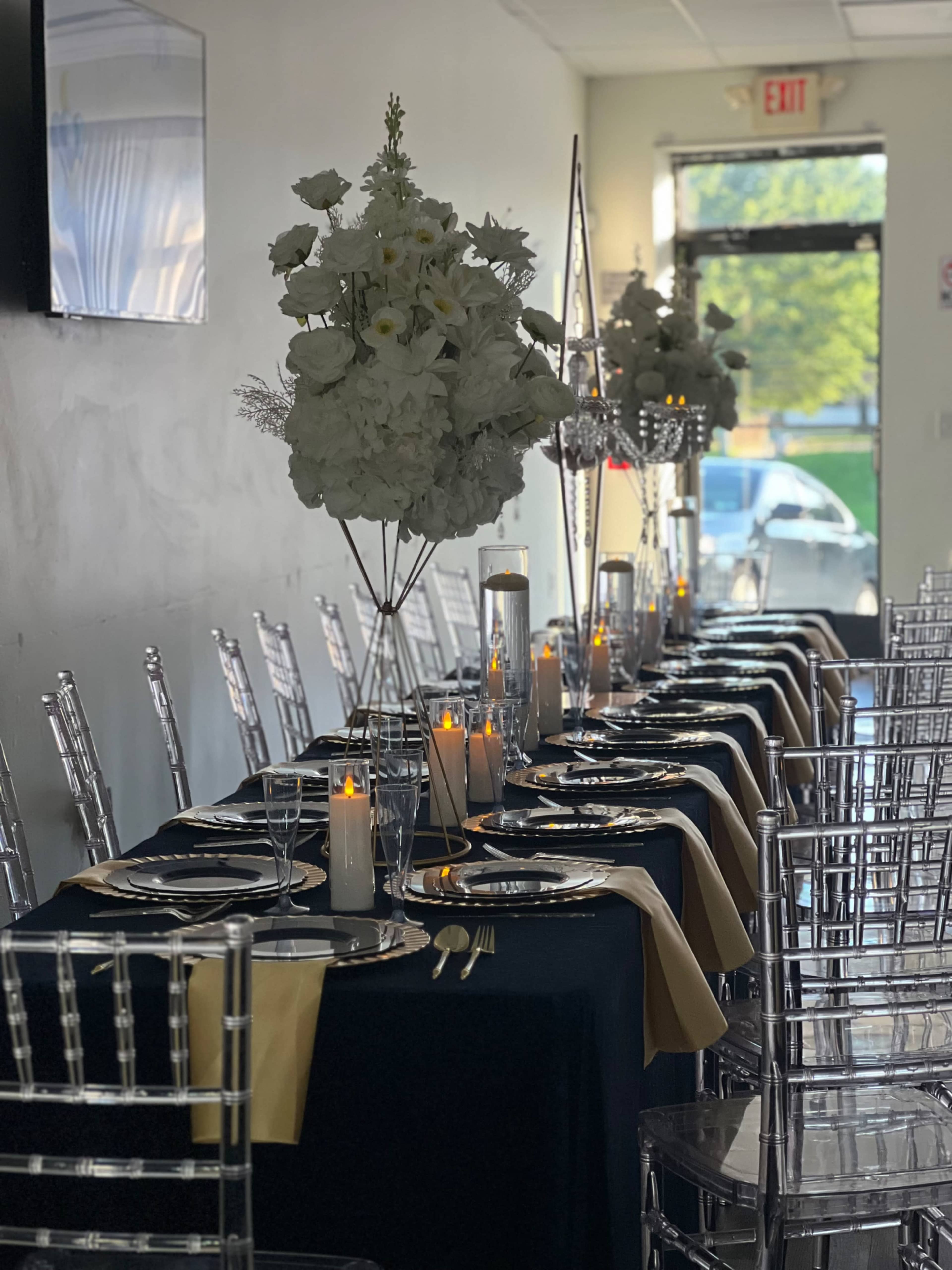 A long banquet table is elegantly set with black and gold table linens, floral centerpieces, candles, and clear acrylic chairs in a well-lit room.