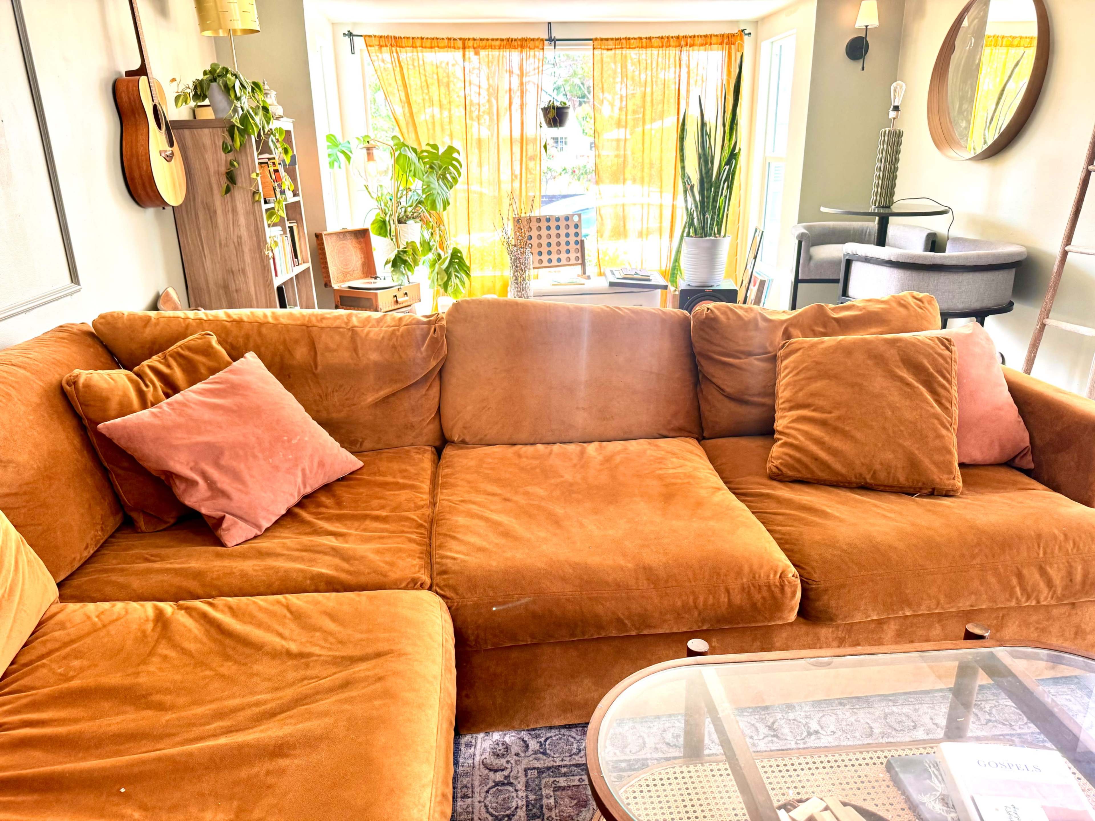 A spacious living room features an orange couch with several pillows, complemented by a guitar on the wall and plants near large windows.