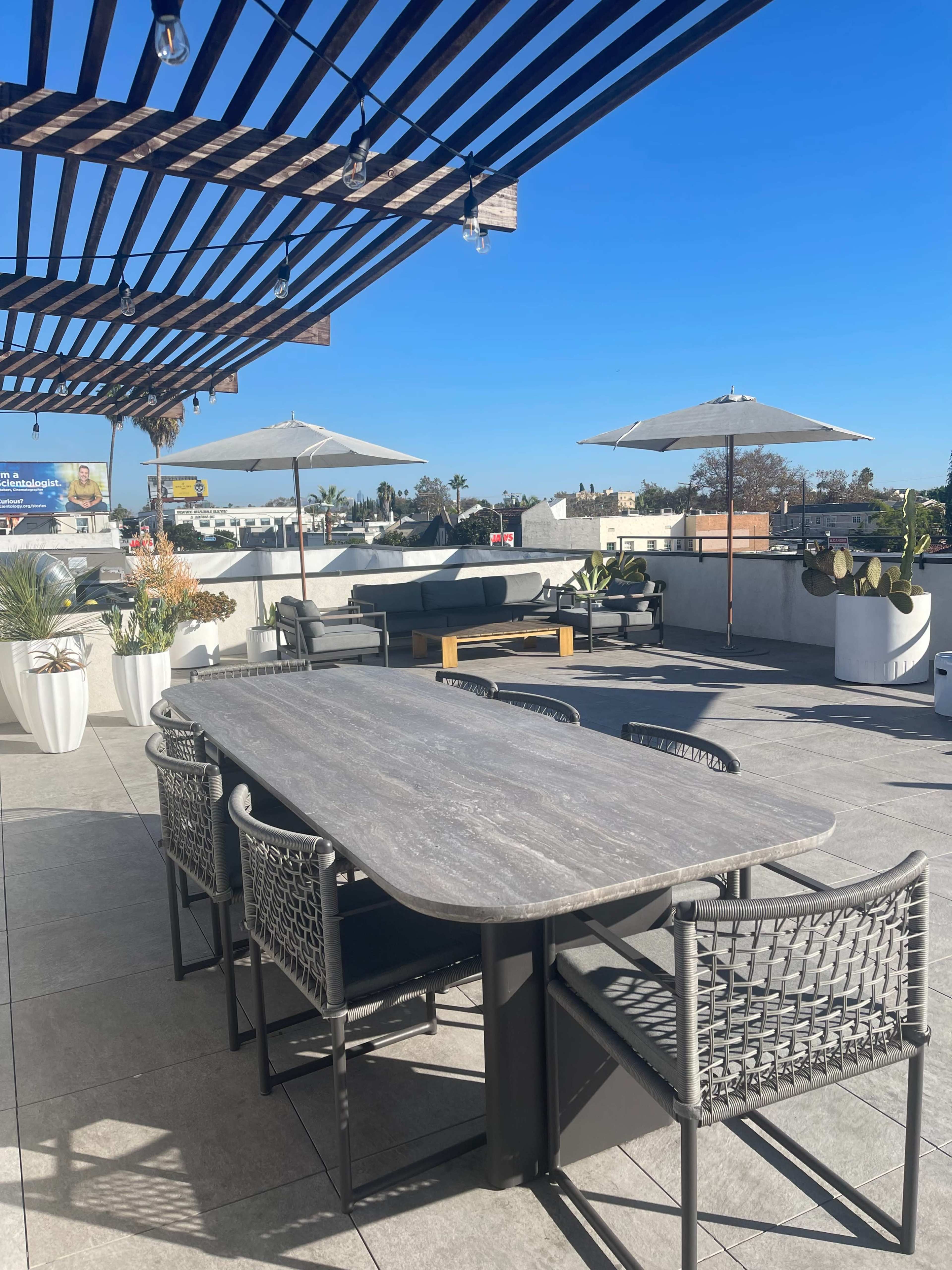 The image shows a rooftop terrace featuring a large table with chairs, several umbrellas, and lounge seating, all under a wooden pergola against a clear blue sky.