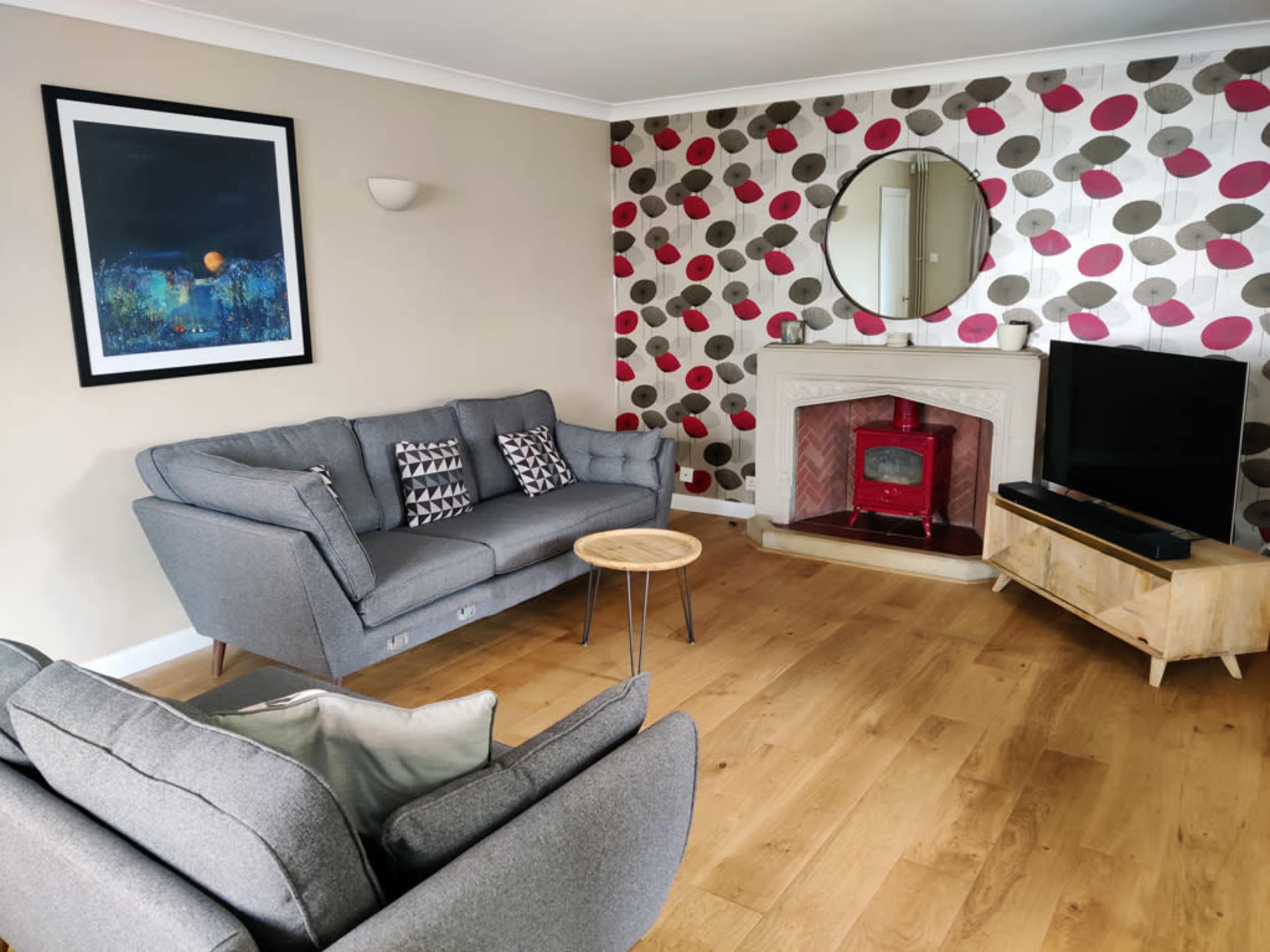 The living room features a gray sofa set, a round mirror above a fireplace, and a television mounted on a wooden cabinet against a wall decorated with patterned circles.