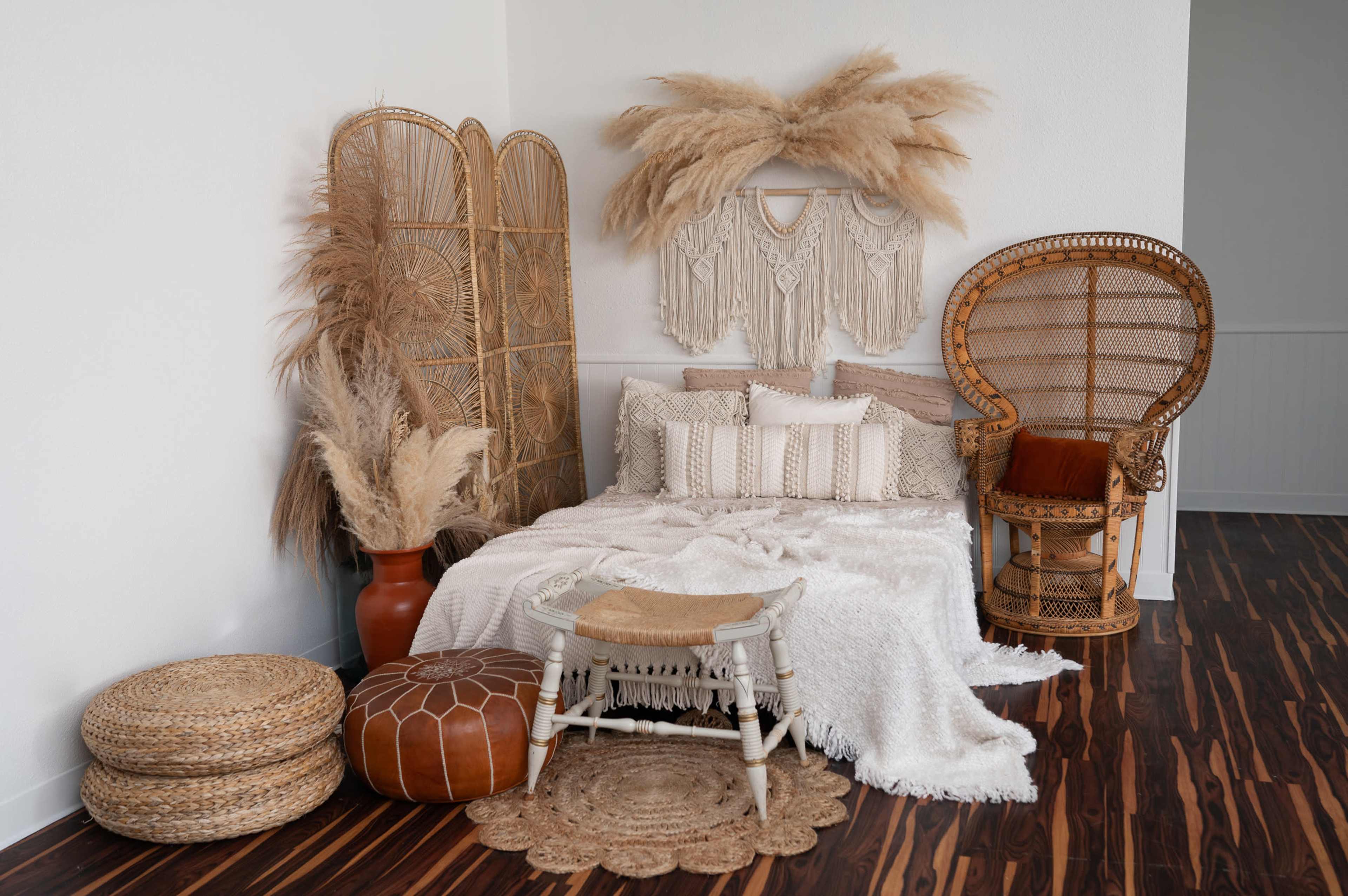 The image shows a cozy bedroom corner decorated with a bed draped in a white blanket, surrounded by woven furniture, pampas grass, and decorative baskets.