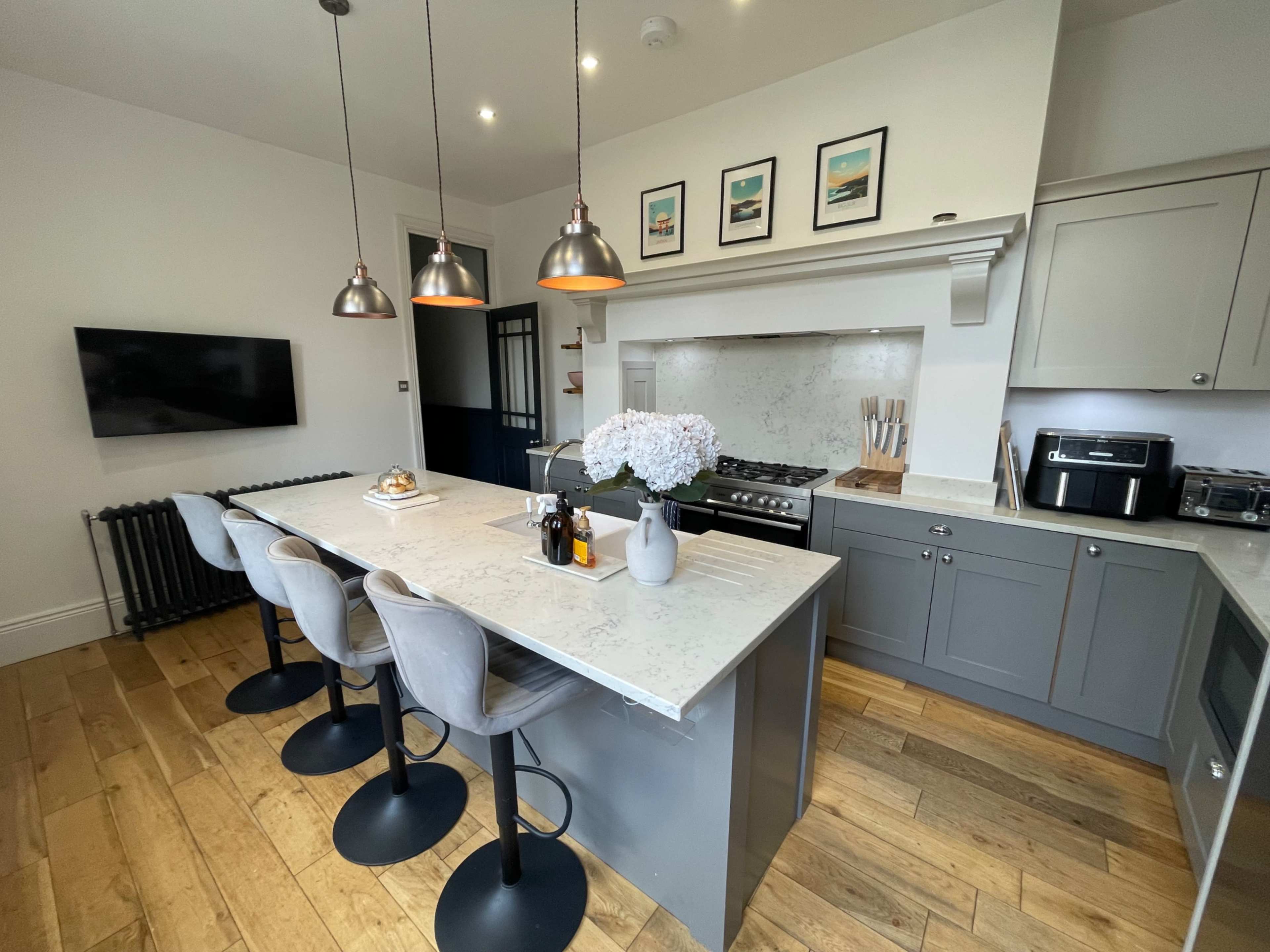 A modern kitchen features a central island with four stools, pendant lighting, and a wall-mounted television, complemented by gray cabinetry and a wooden floor.
