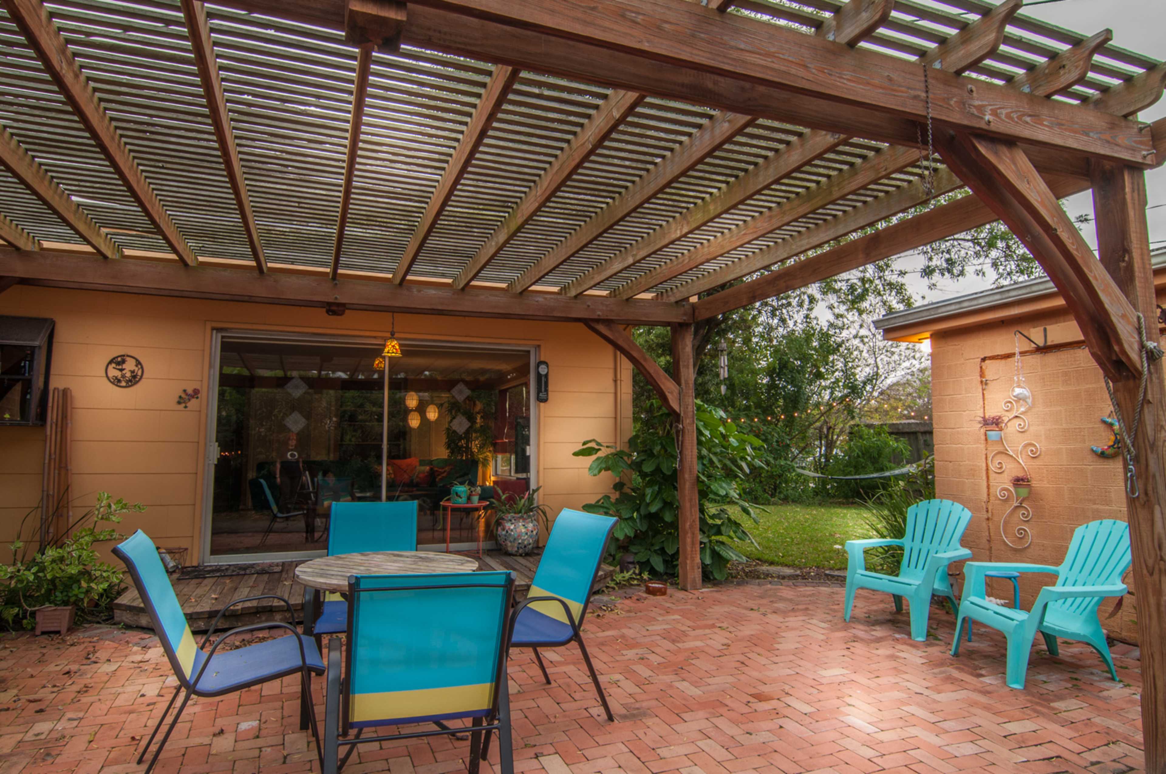 A covered patio features a circular table surrounded by six chairs and two lounge chairs, set against a backdrop of a well-maintained garden.