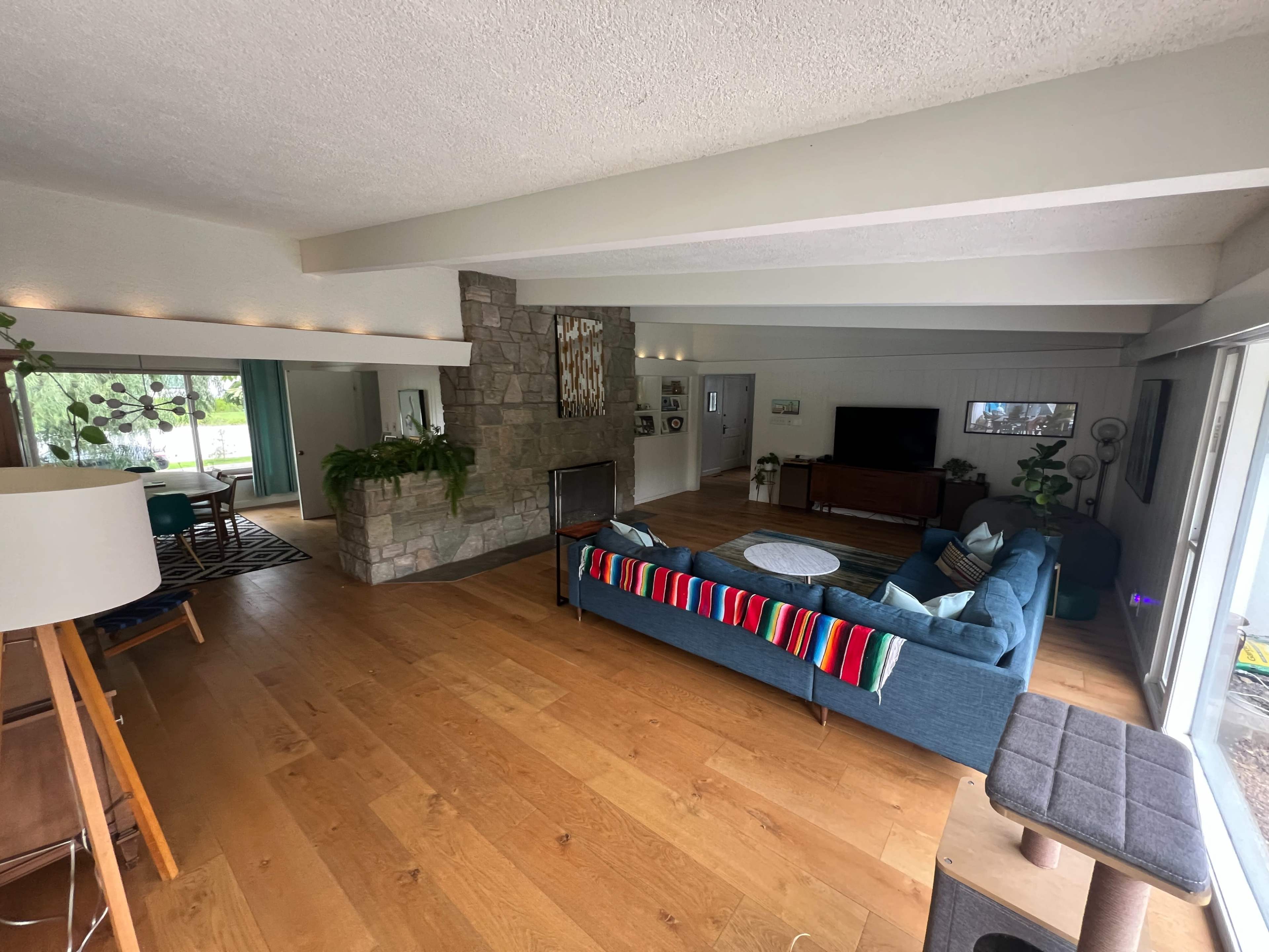 The image shows a spacious living room with wooden flooring, a stone fireplace, a blue couch, and large windows providing natural light.