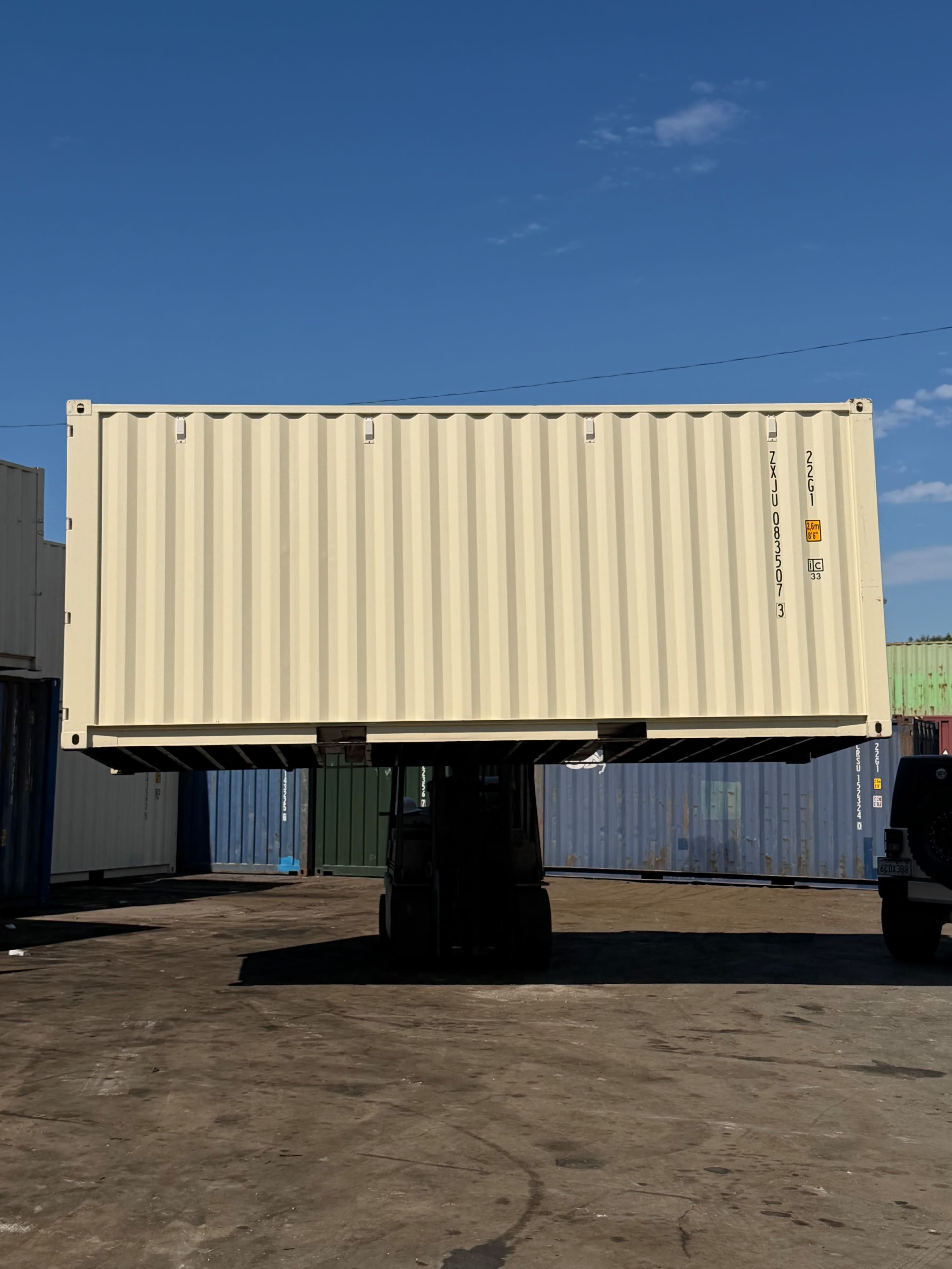A forklift holds a large shipping container above ground in a storage yard surrounded by other containers.