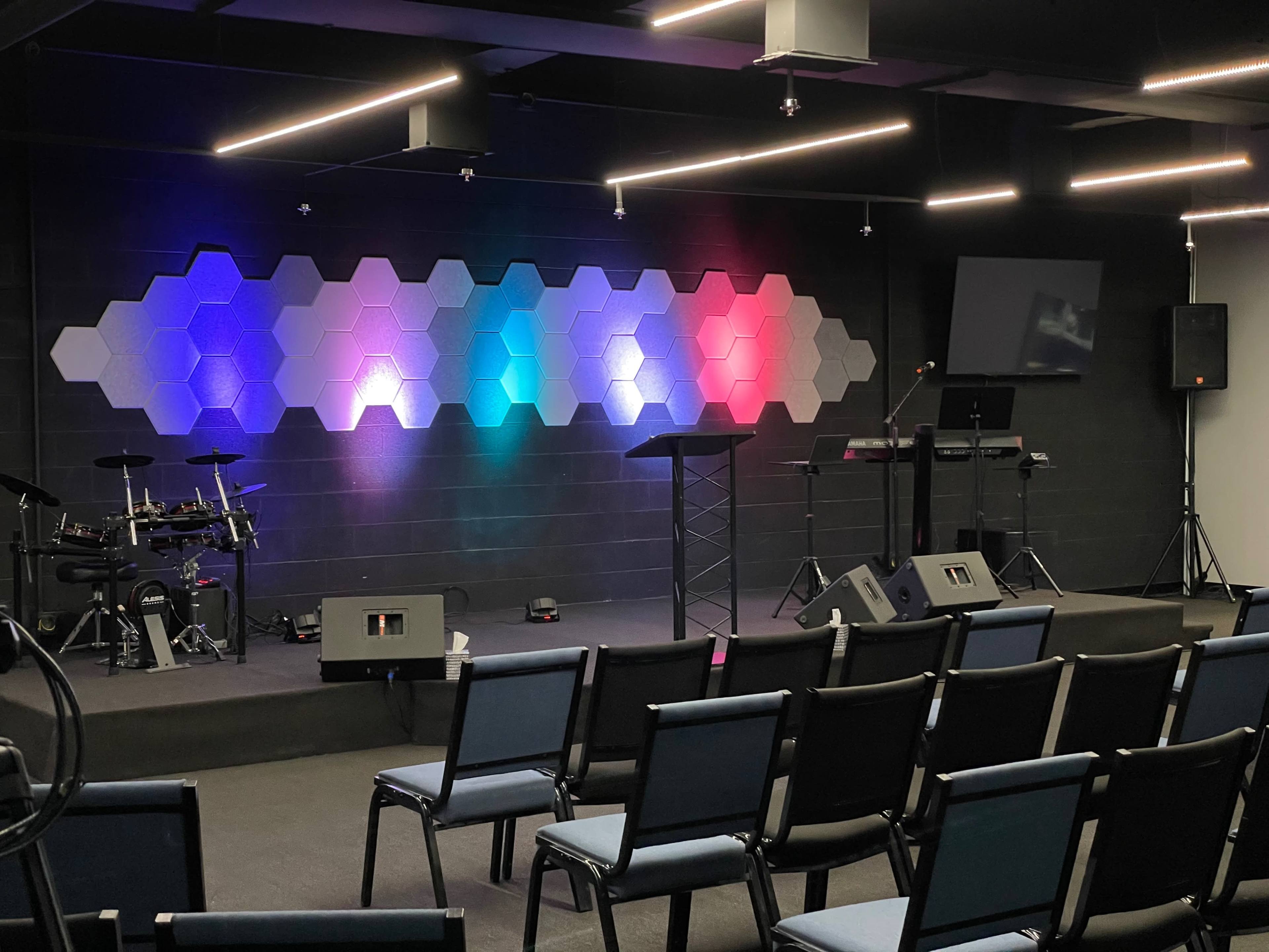 A modern stage setup features a hexagon-patterned backdrop with colored lights, a drum kit on one side, and seating arranged in front.