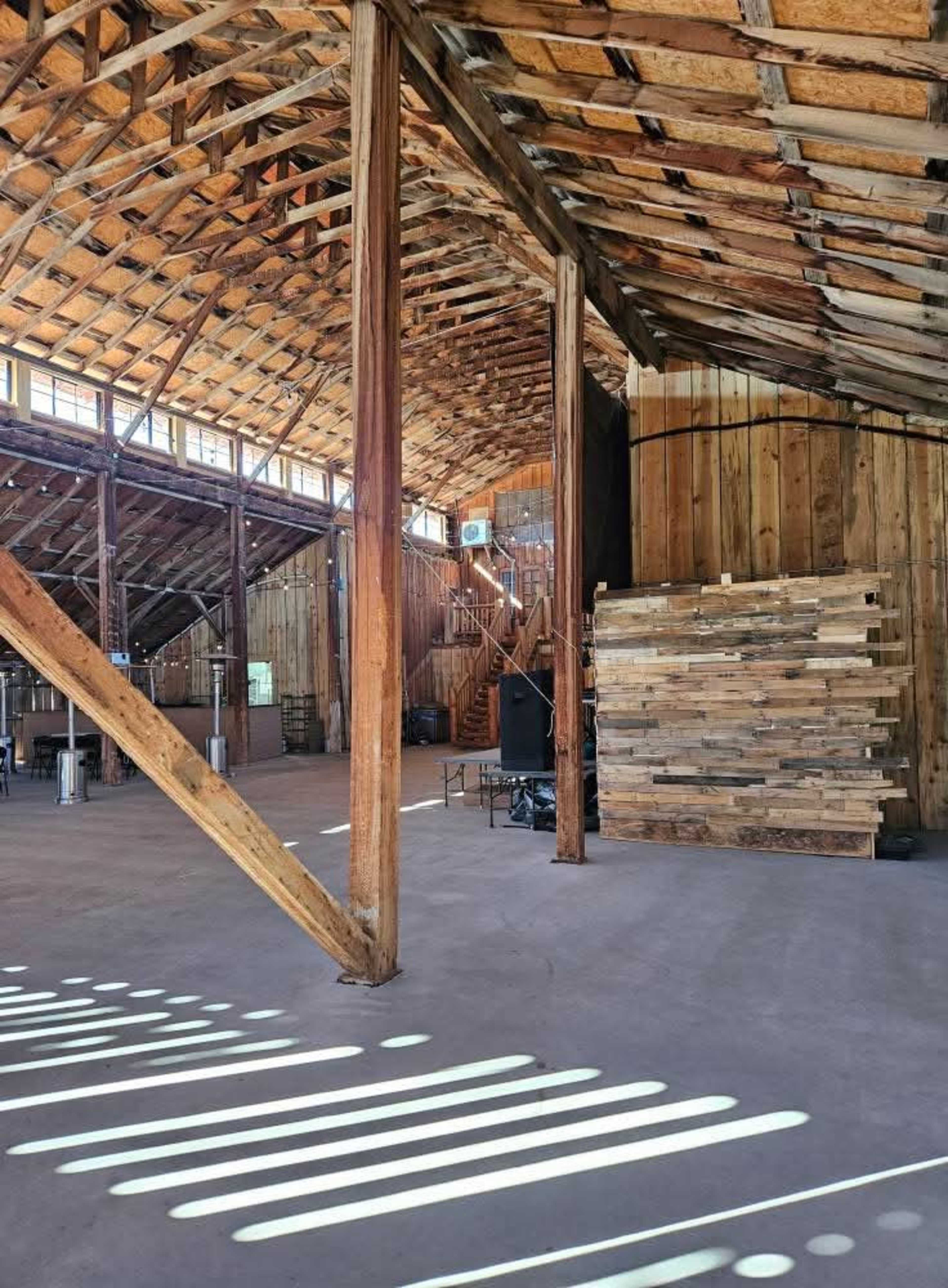 The interior of a wooden barn features exposed beams, a loft area, and stacks of lumber along the wall.