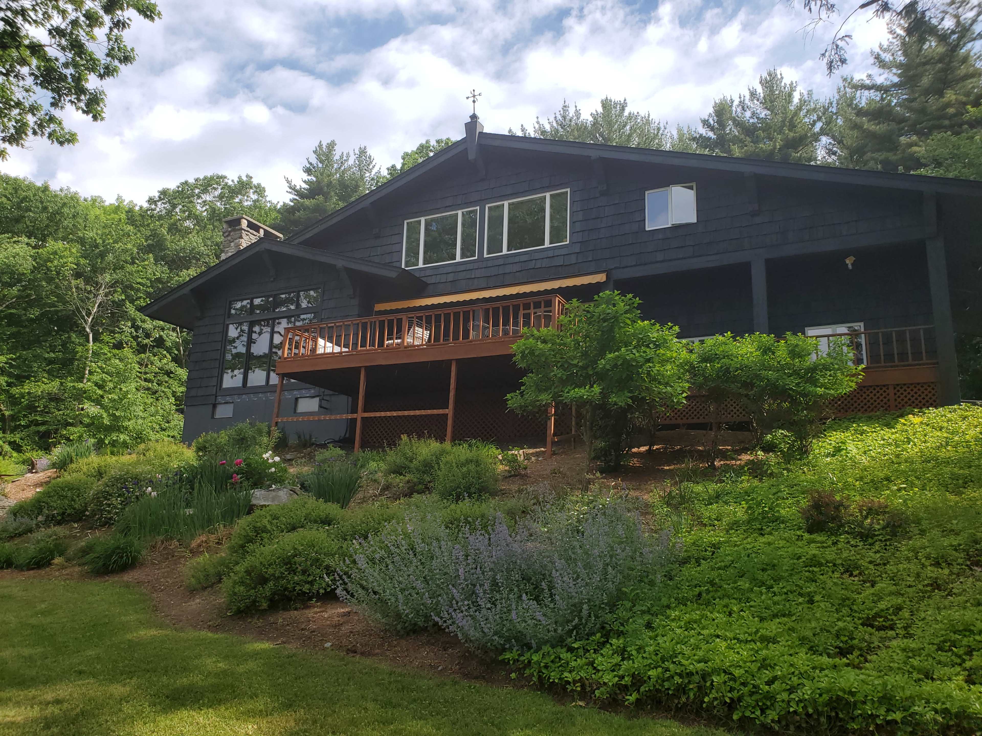 A large dark-colored house with multiple windows and a wooden deck is situated on a landscaped yard featuring various shrubs and flowers.