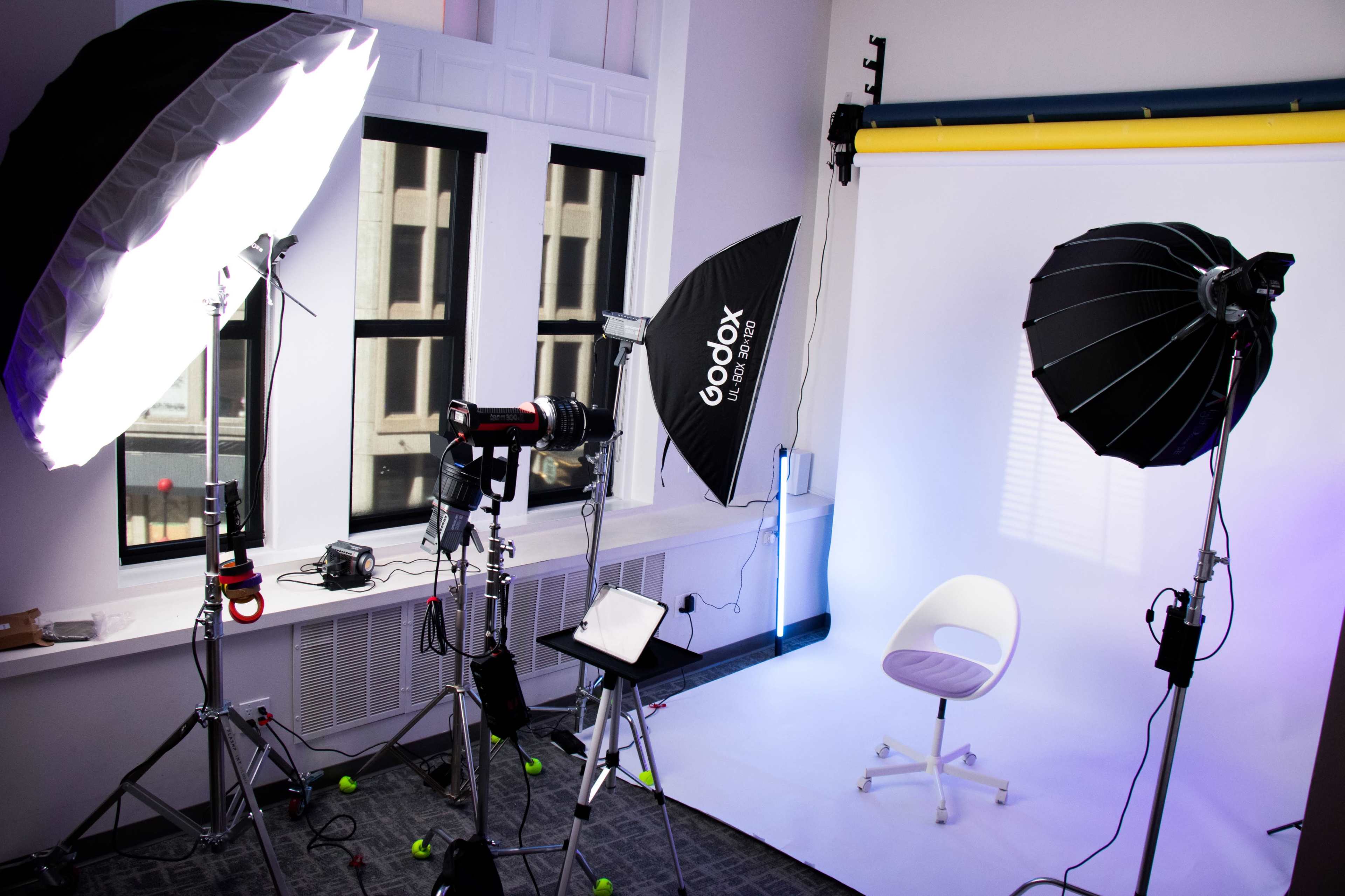 The image shows a photography studio set up with multiple softbox lights, a camera on a tripod, and a white backdrop with a lone chair in the center.