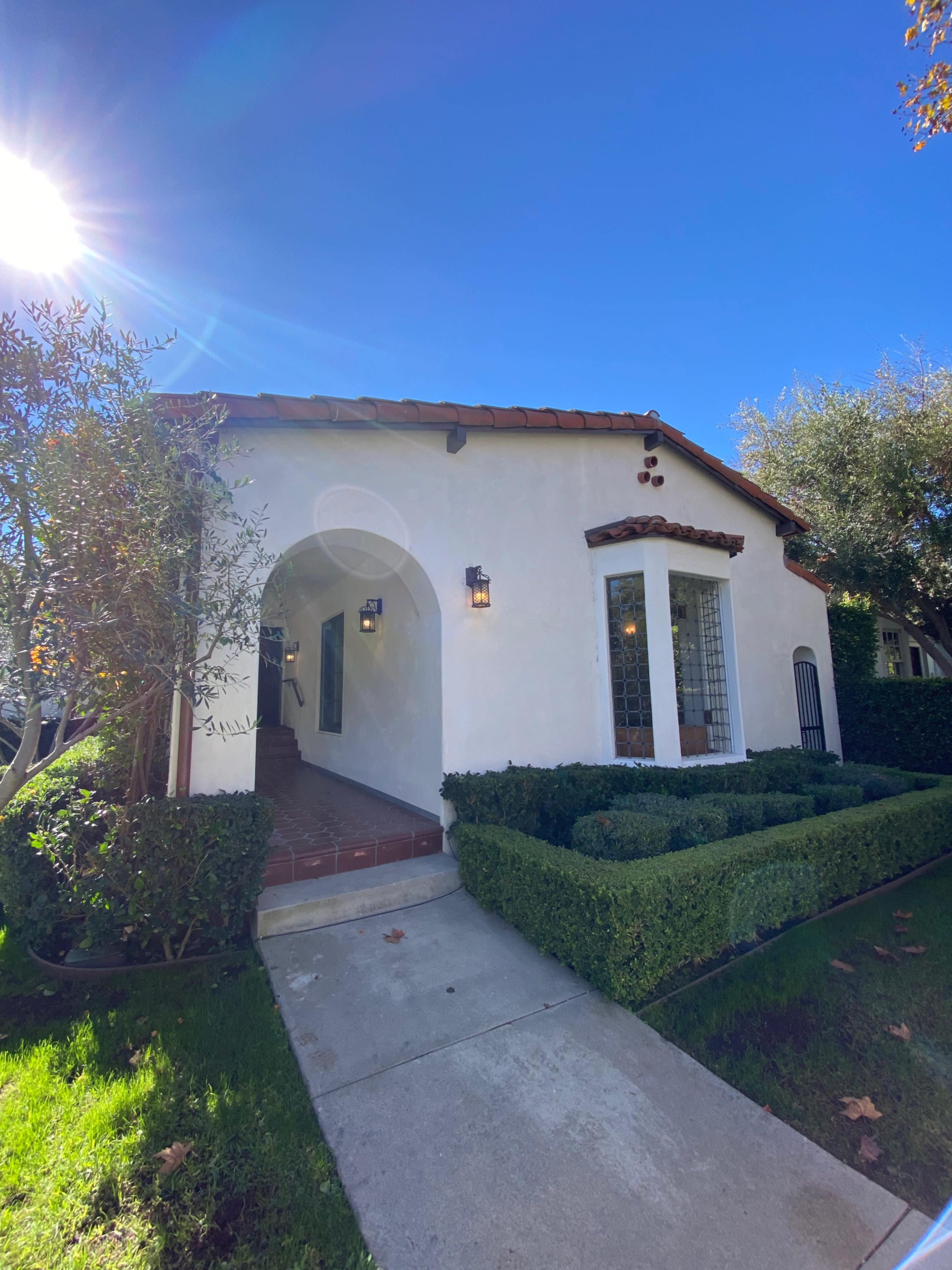 A white stucco house with a red-tiled roof features a landscaped lawn and a concrete walkway leading to an arched entrance.