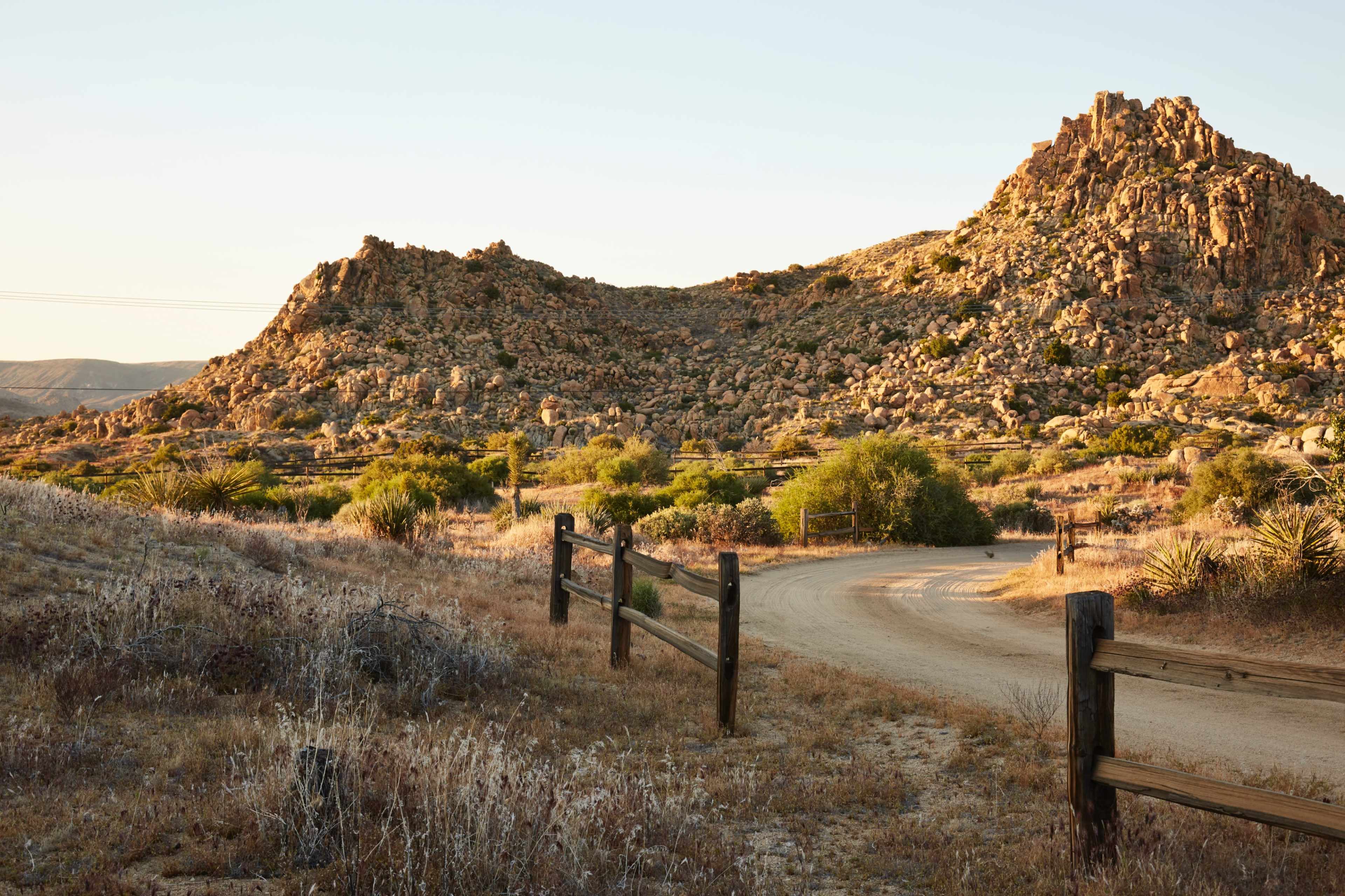 A dirt road winds through a rocky landscape with scattered shrubs and a fence running alongside it.