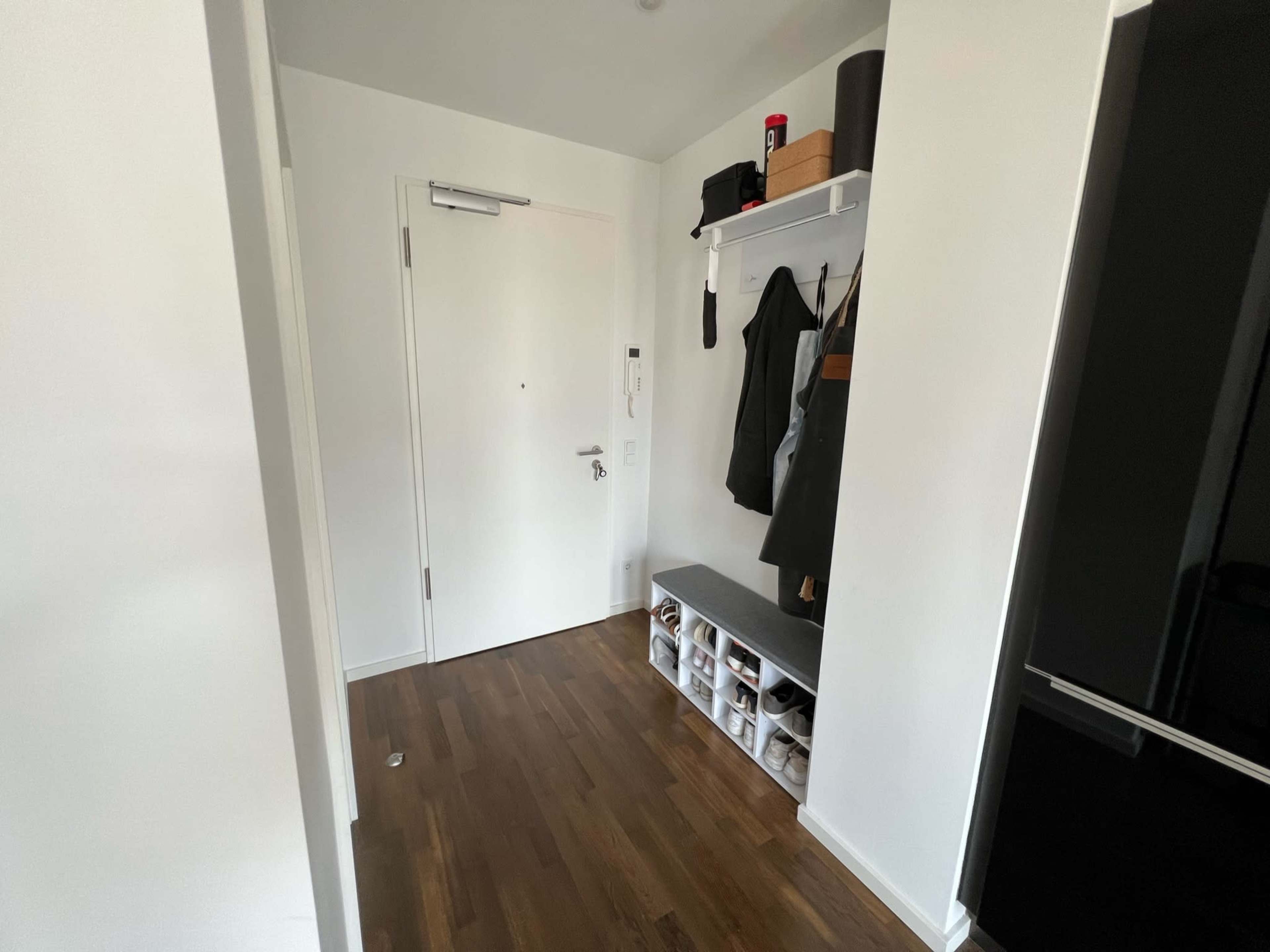 A hallway with a door on one side, a shoe organizer along the wall, and a shelf holding bags and personal items above.