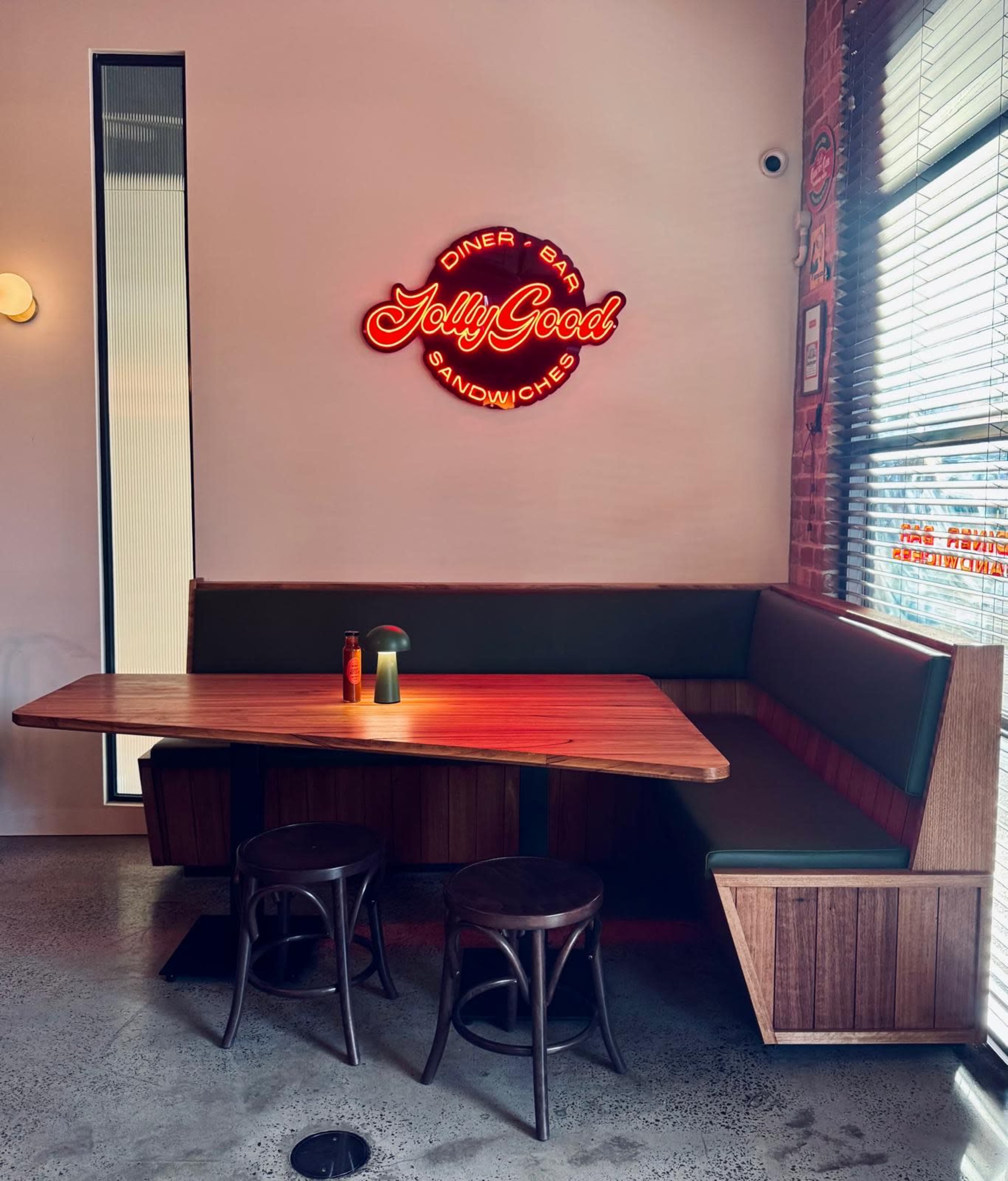 The image shows a modern diner interior with a wooden table, chairs, and a neon sign that reads "Jolly Good Sandwiches."
