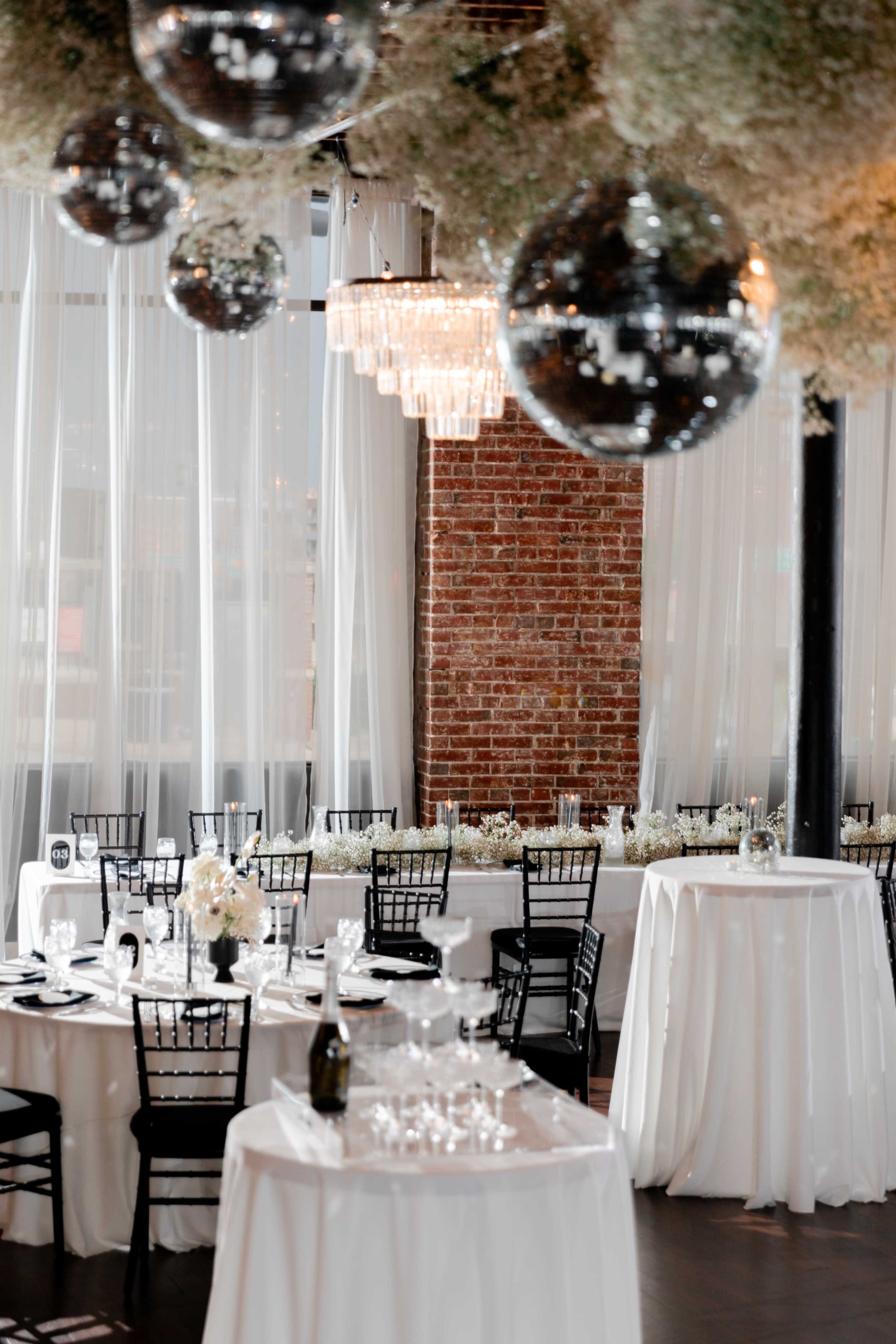 The image shows an elegantly decorated indoor event space featuring round dining tables with white tablecloths, black chairs, and hanging disco balls amidst floral arrangements.