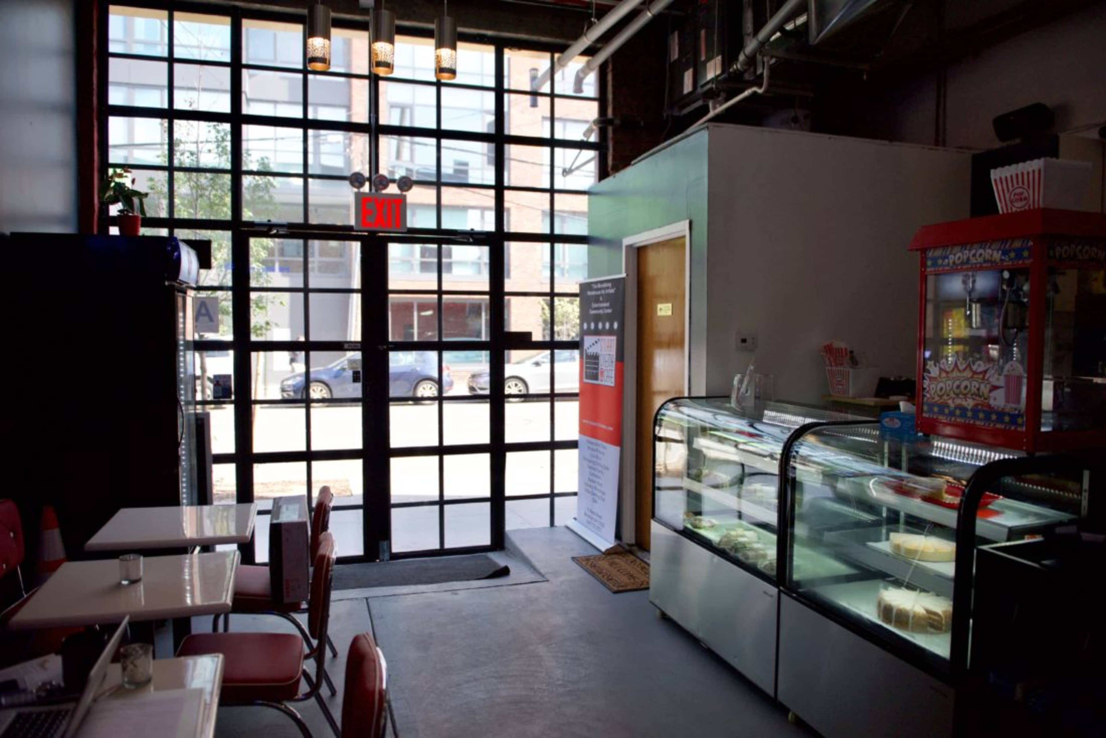 A café interior with large windows, a glass display case for desserts, and red chairs at small tables facing the entrance.